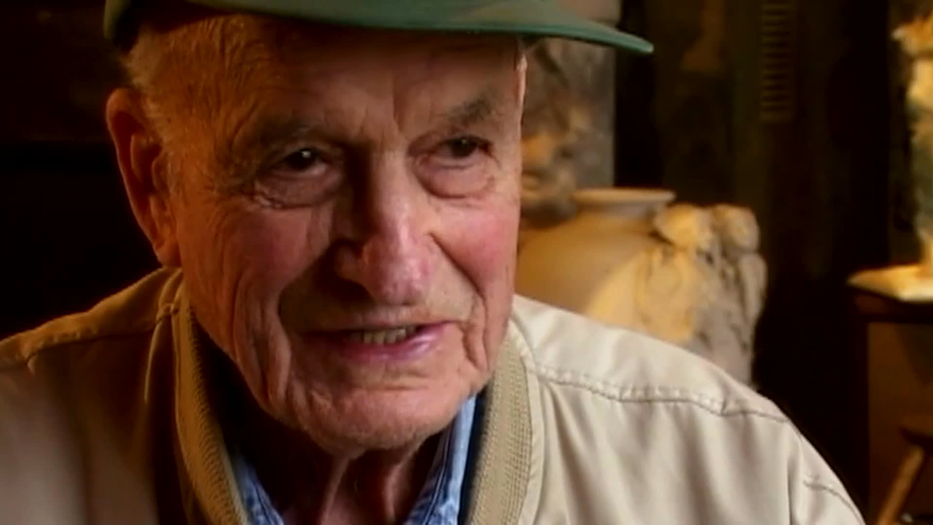 An elderly art collector describes how buying art gradually filled his home and a storehouse, leading to the construction of another building. He speaks in a warmly lit room among artworks, while outdoor views show a large structure under construction.