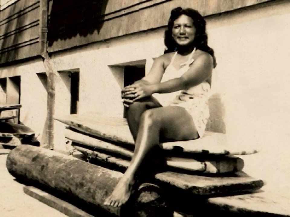 Performers in traditional attire engage in a hula dance on a tropical lawn before transitioning to vintage home movie footage of a couple sharing lighthearted moments in a garden setting.