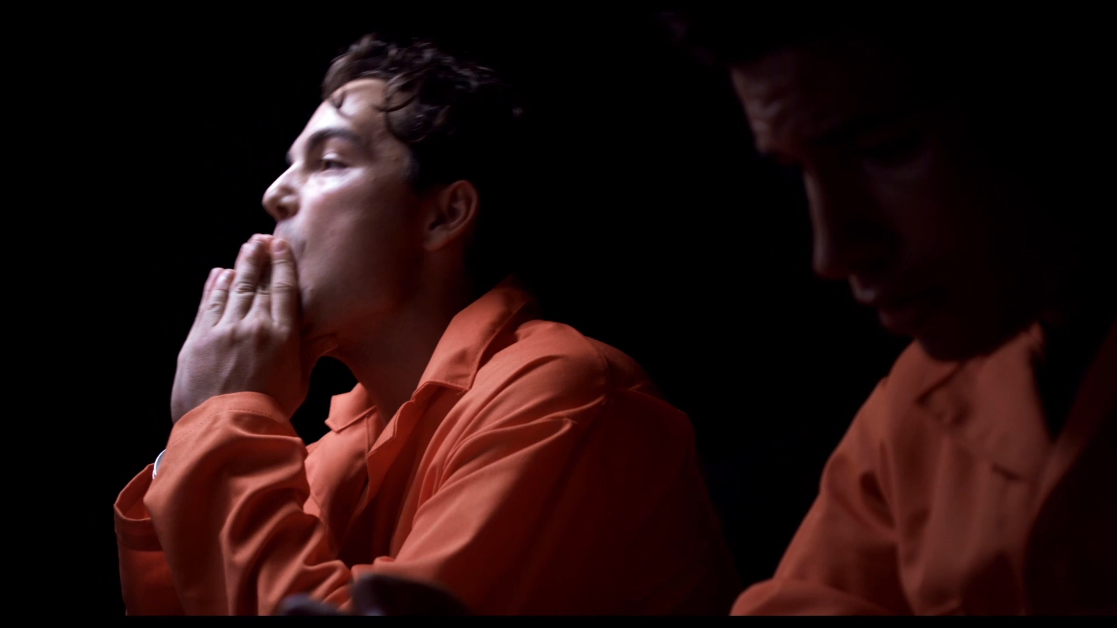 Two individuals in orange jumpsuits engage in a hushed conversation while handcuffed in a dimly lit environment. A walkie-talkie sits nearby as one participant appears distressed and reflects on their situation.