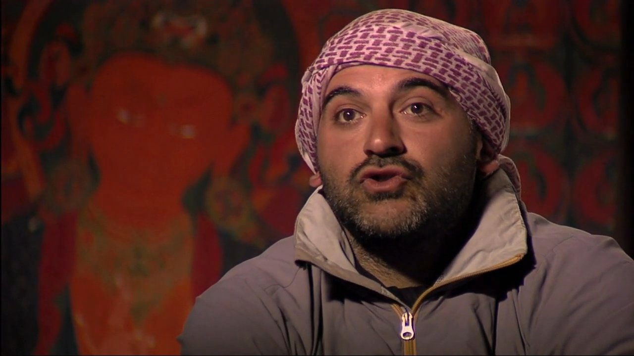 A man in traditional headwear discusses cultural experiences during a candid interview, contrasted with exterior shots of a monastery and bustling crowds in a nearby village.