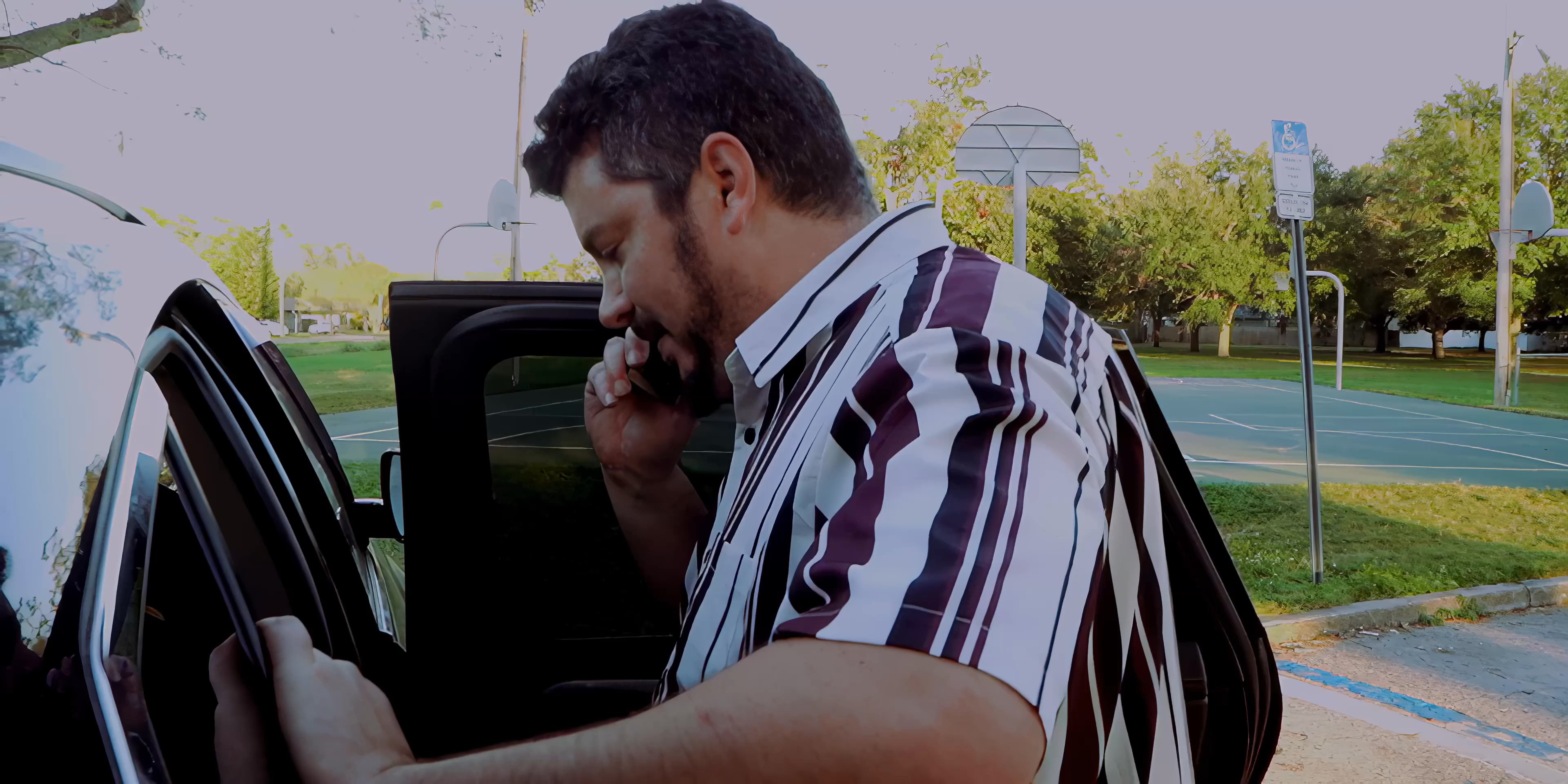 A man in a striped shirt engages in a mobile phone conversation while entering a parked vehicle in an outdoor setting.