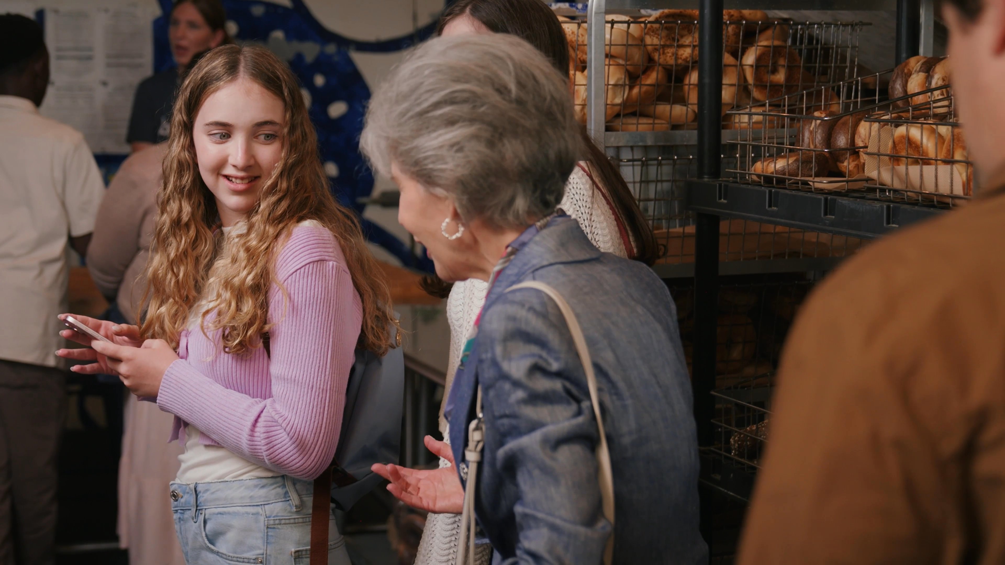 A group of diverse individuals engages in a conversation at a bakery counter, with an older woman appearing to guide or advise a younger woman.