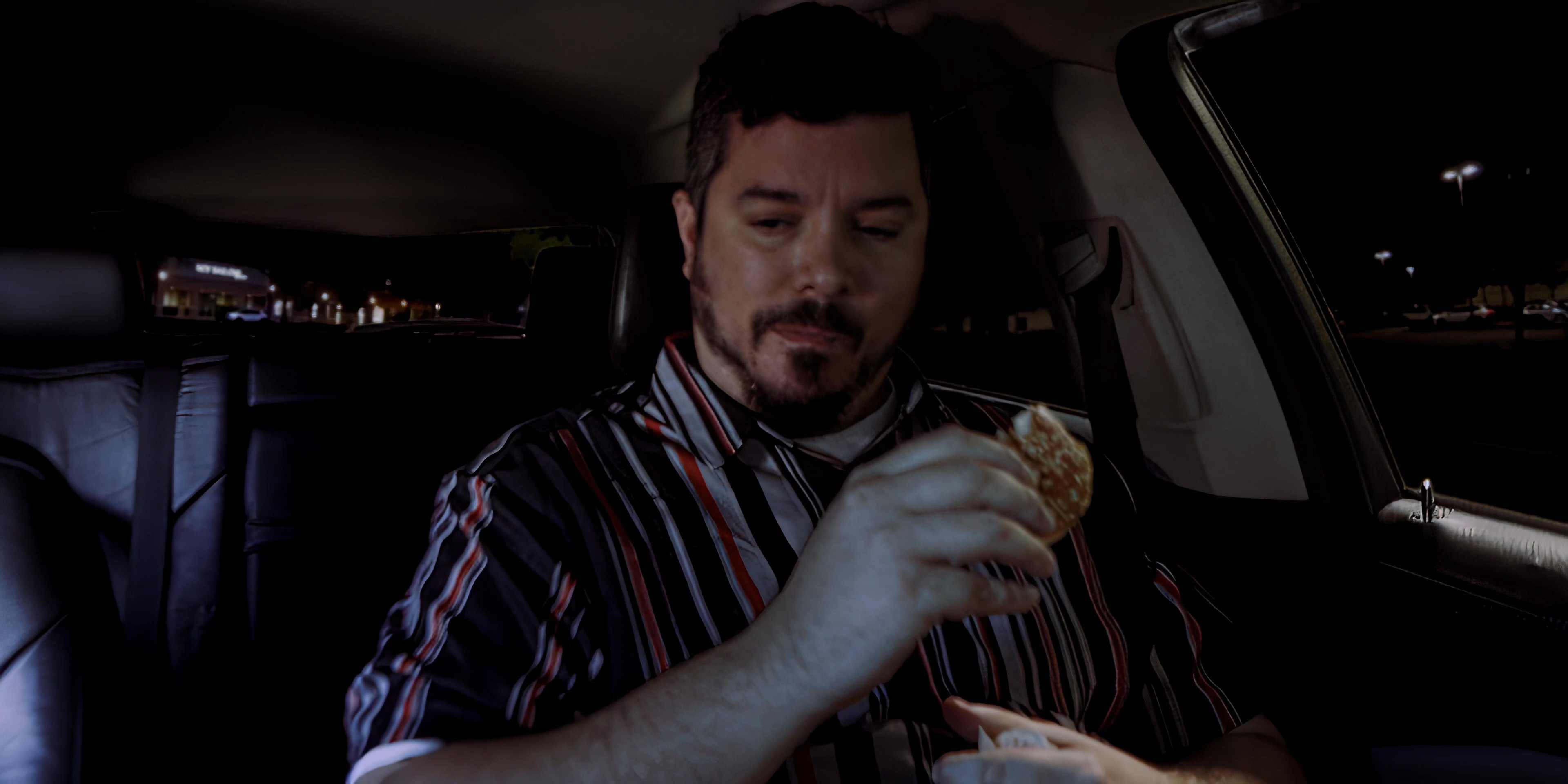 A man sits in the passenger seat of a vehicle at night and consumes a hamburger.