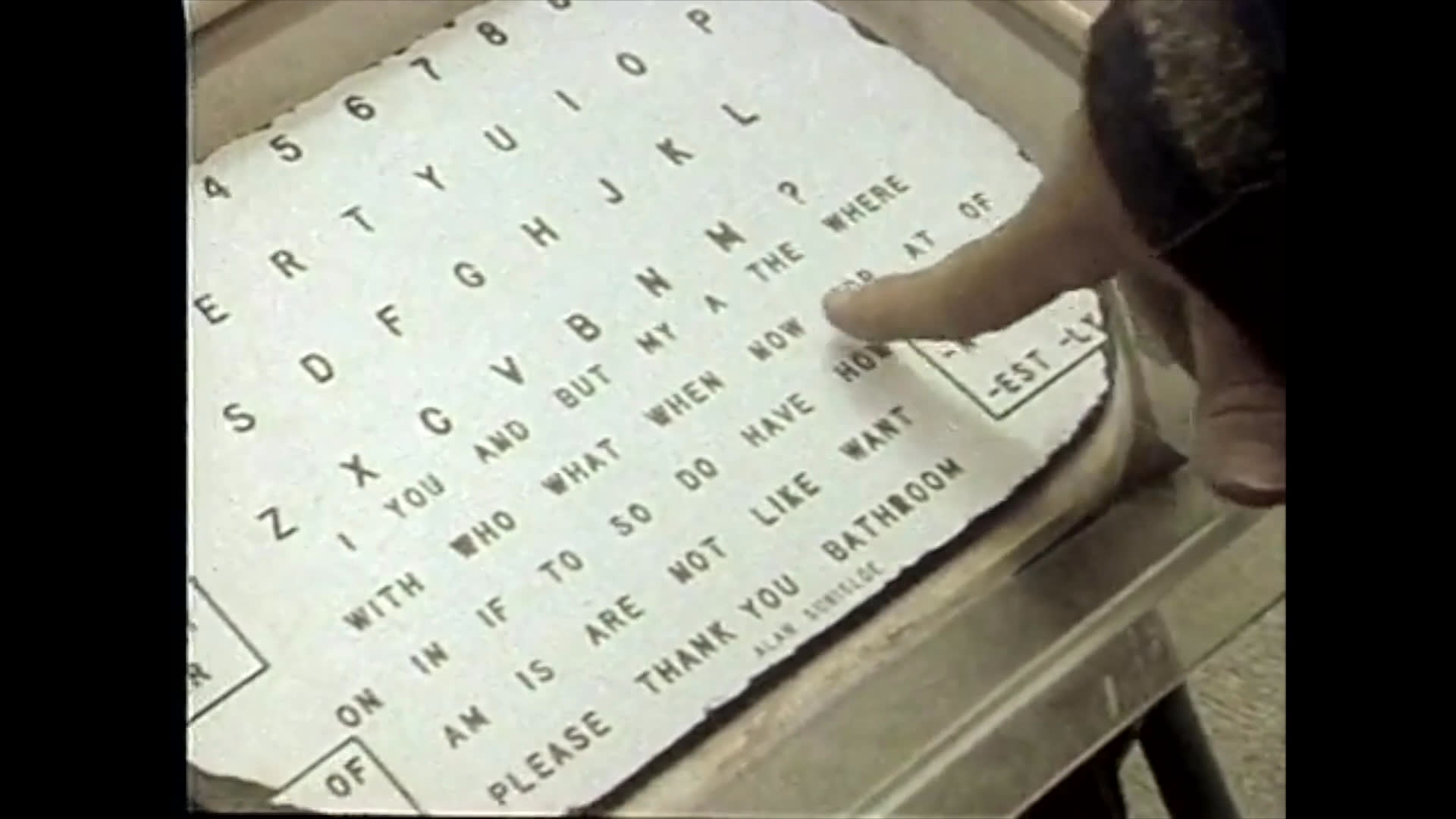 A man guides a person using a mobile communication board, demonstrating its use for expressive language in an outdoor park setting.