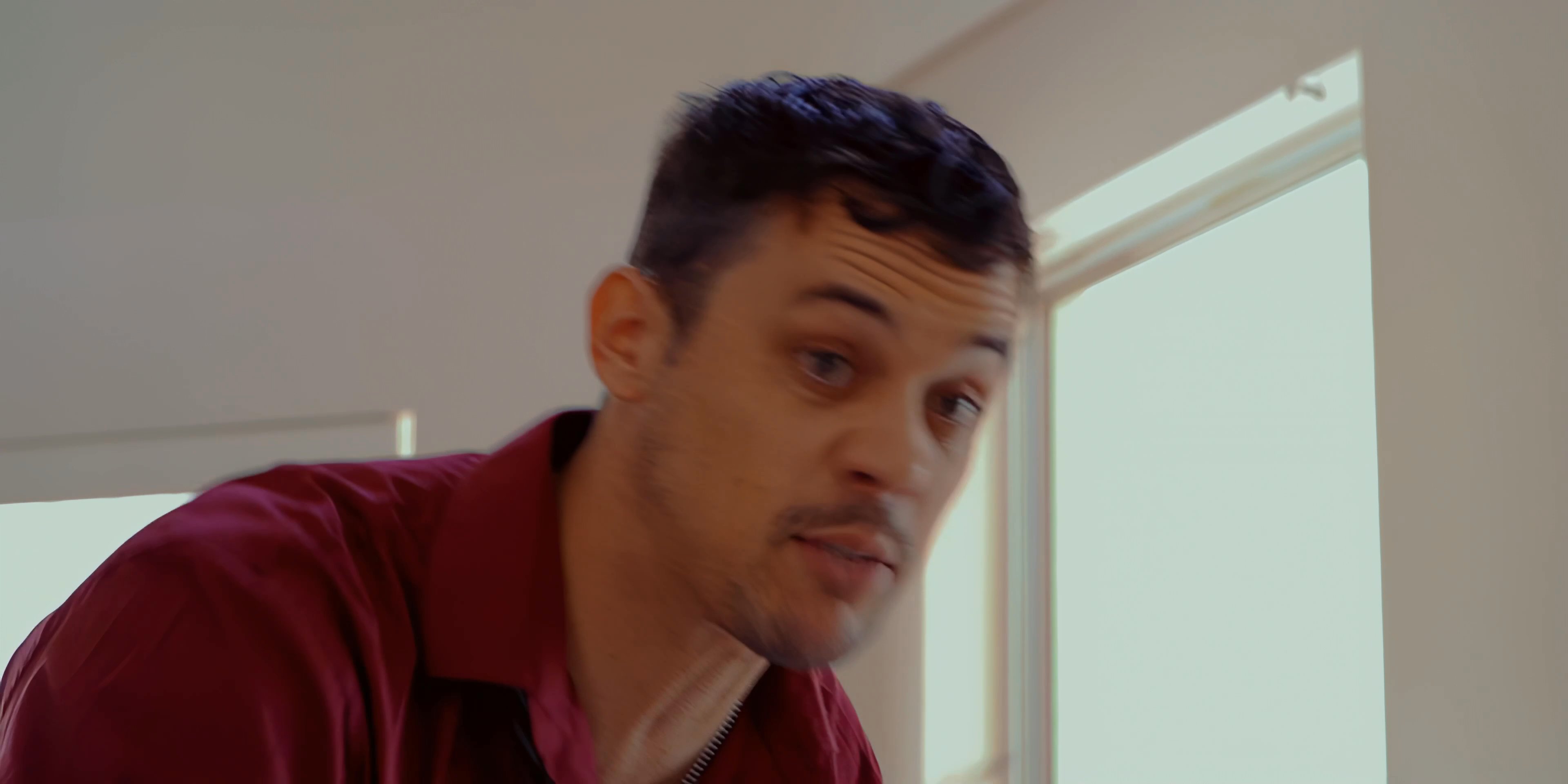 A man in a red shirt expresses intense frustration while arguing with an older man in a suit who sits on a bed. Their heated exchange suggests a high-stakes interpersonal conflict.