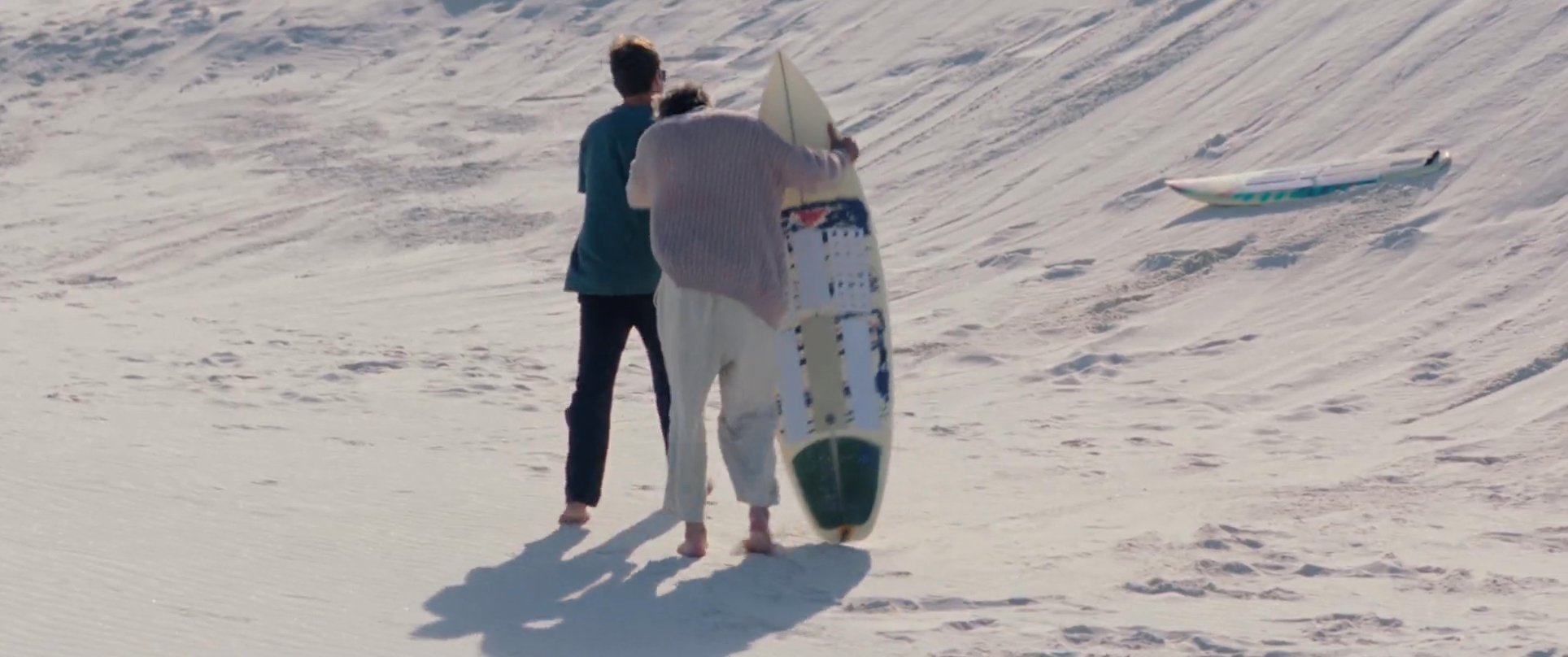 Two individuals prepare for sandboarding on expansive white sand dunes before one descends the slope on a board.