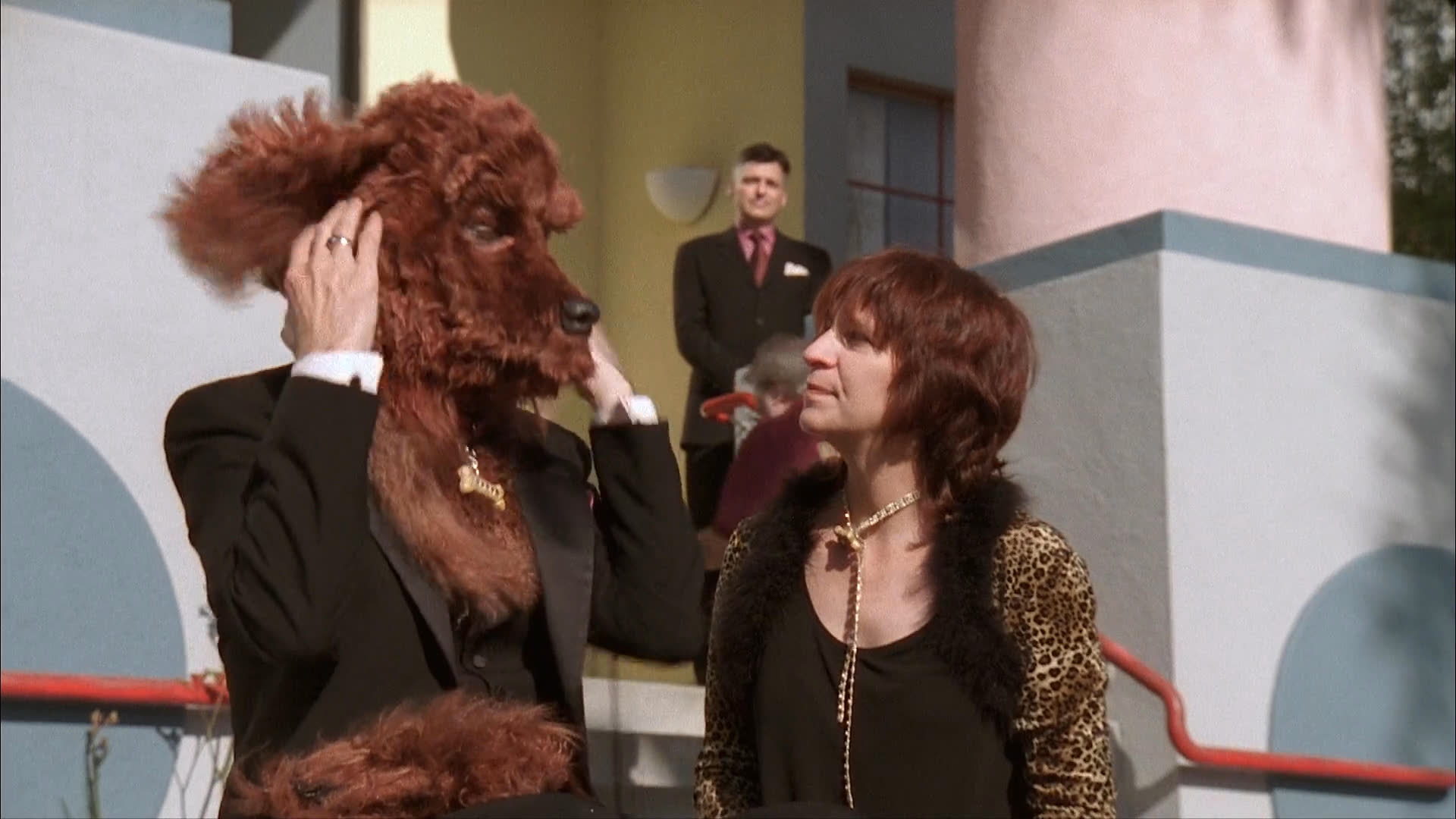 A man wearing a large brown dog costume interacts with a woman in a leopard-print jacket outdoors while others observe in the background.