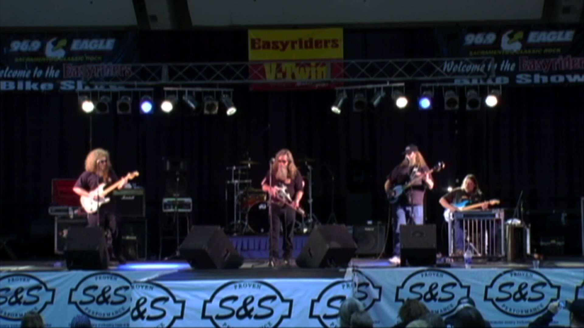 A rock band performs on a concert stage with the lead singer and guitarist in view. The musicians play their instruments and sing to an audience during a live music event.