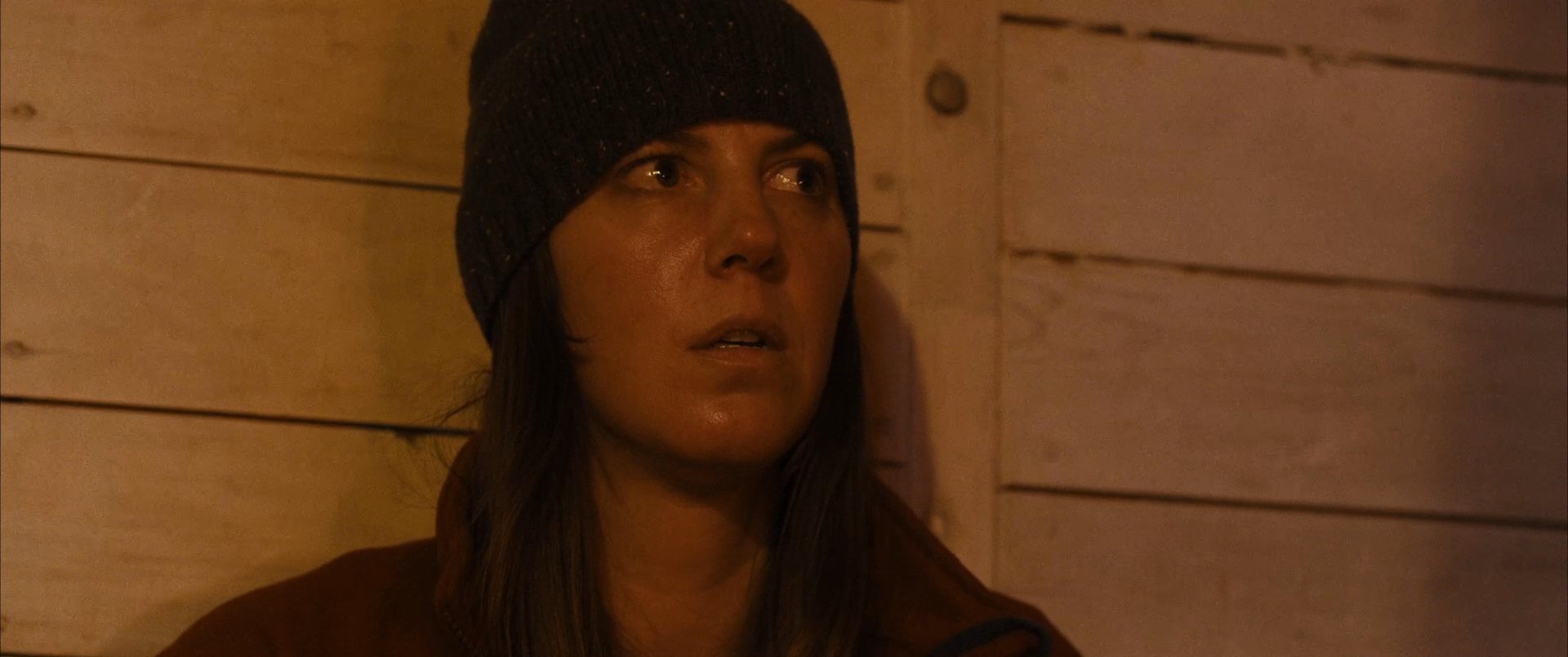 Two individuals engage in a somber conversation within a dimly lit, candlelit cabin setting. The exchange maintains a tense, intimate atmosphere throughout the interaction.