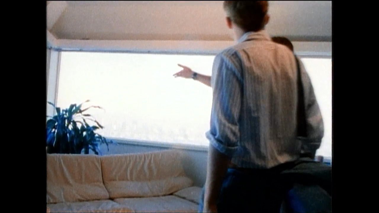 A man and a woman engage in a playful conversation and expressive hand gestures on an outdoor terrace, later transitioning to an indoor living area where they interact with another individual.