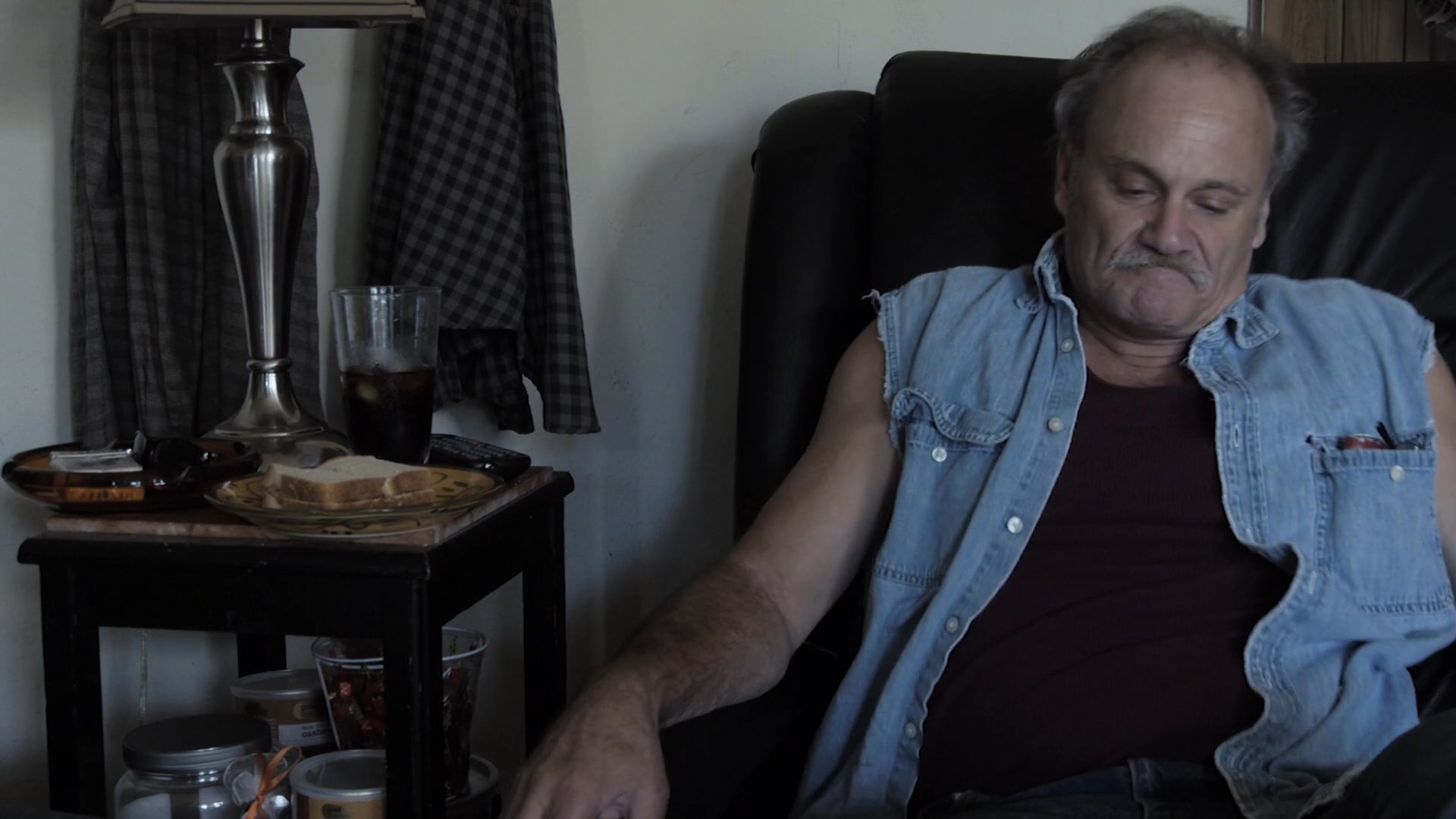 An older man and a young woman sit in a living room, conveying a sense of emotional distance and unspoken tension through their restrained interaction and weary expressions.