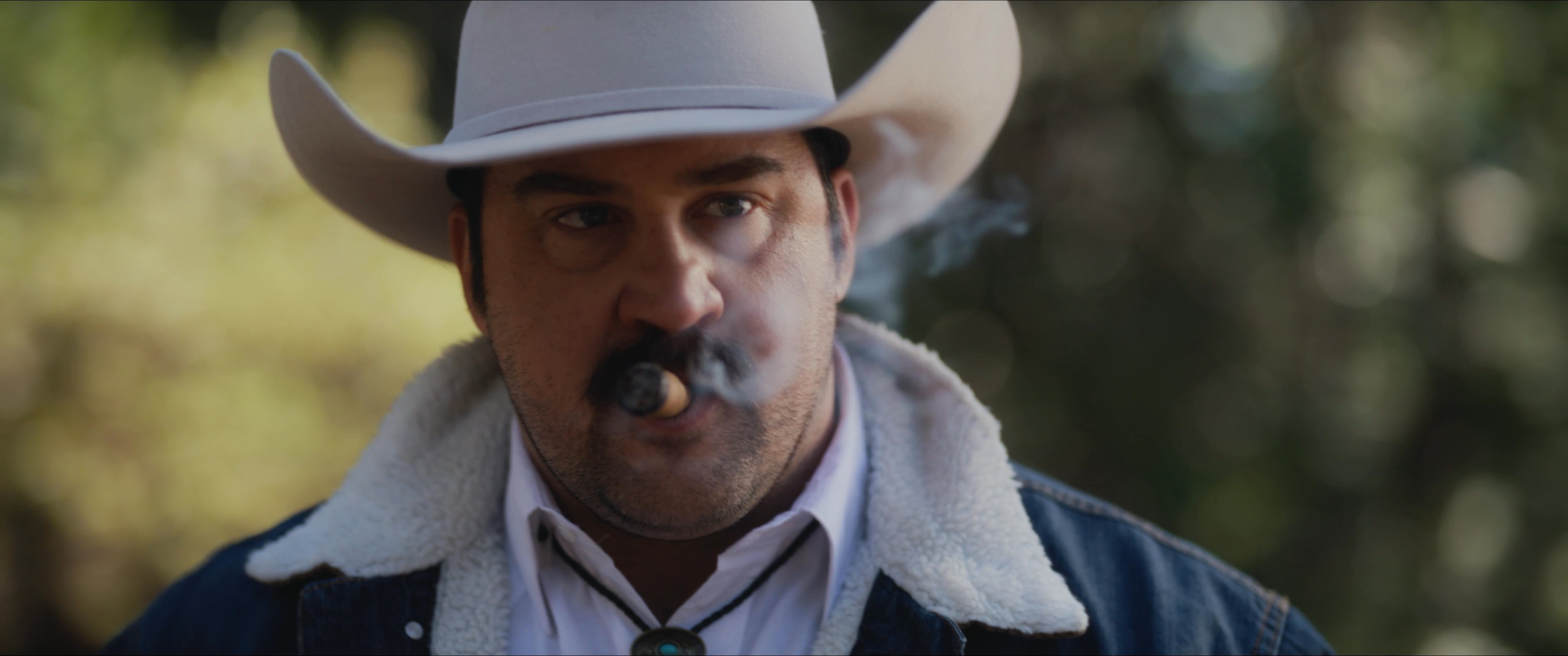 Two men engage in a tense conversation within a wooded environment. One man wears a cowboy hat and smokes a cigar while interacting with a man wearing a tactical cap.
