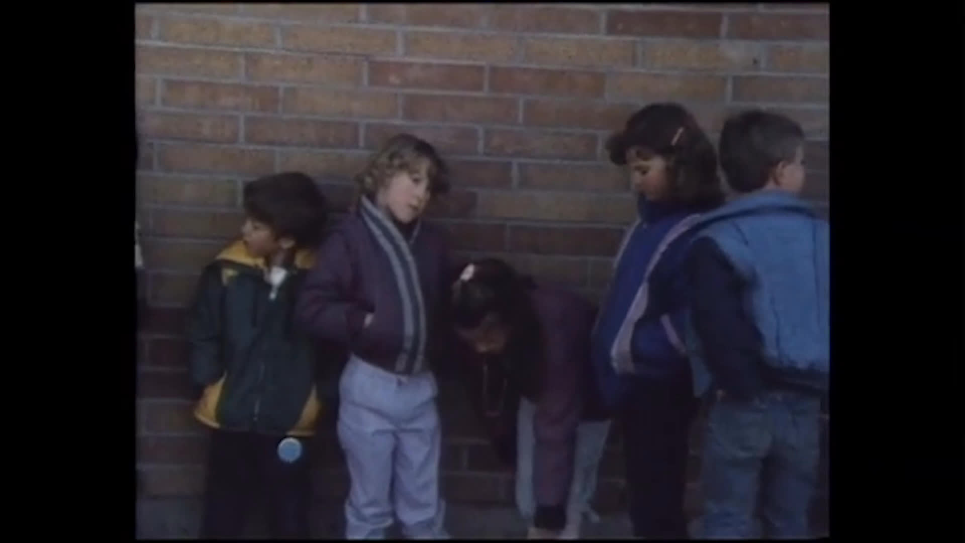 A young girl in red gestures while speaking to a small outdoor gathering, expressing confidence that she can outperform boys at ranch work. Nearby, children run across a yard and gather beside a brick building and basketball hoop.