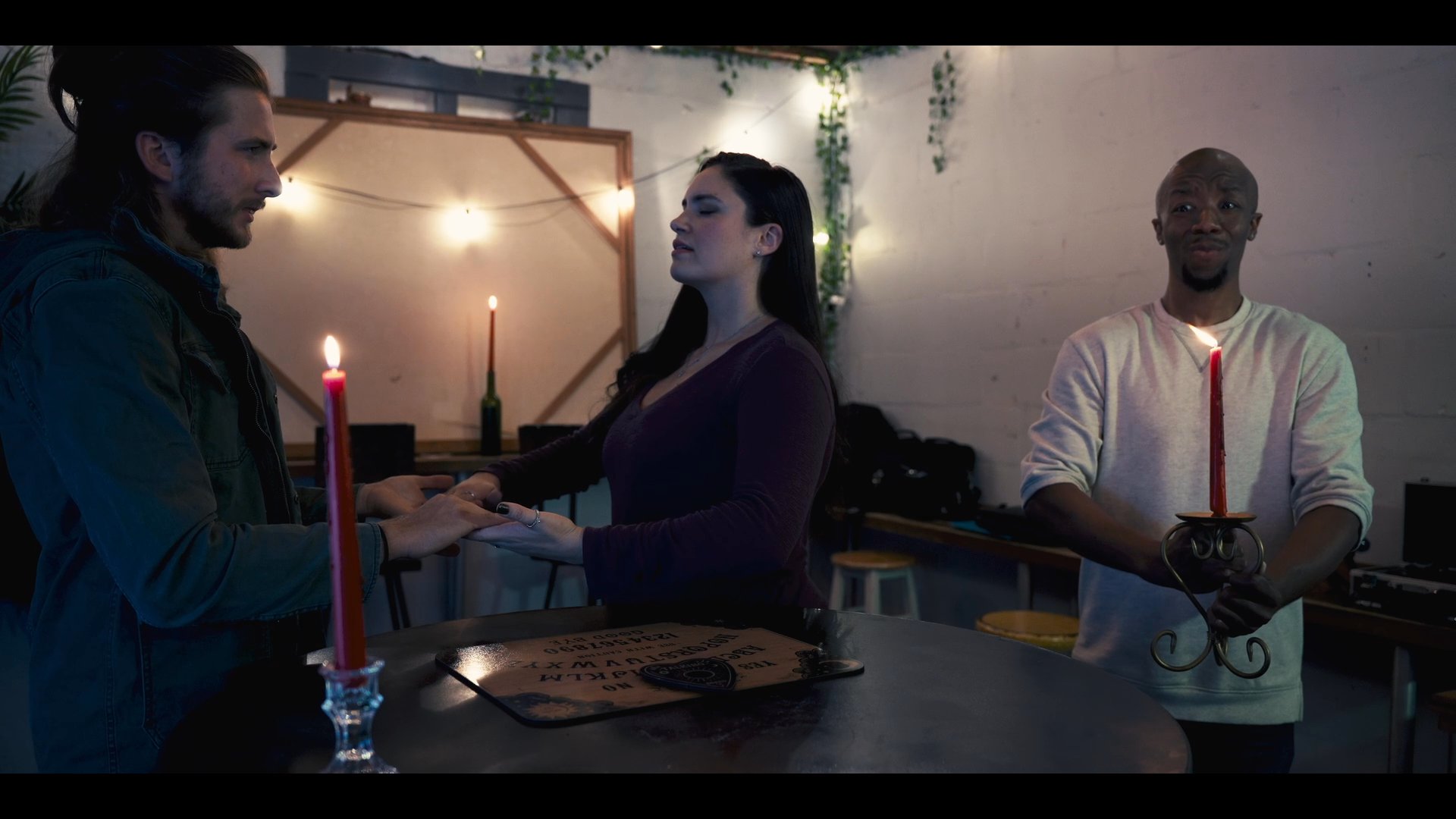 A group conducts a mysterious ritual around a table set with candles and an ouija board, contrasting with a solitary individual resting nearby.