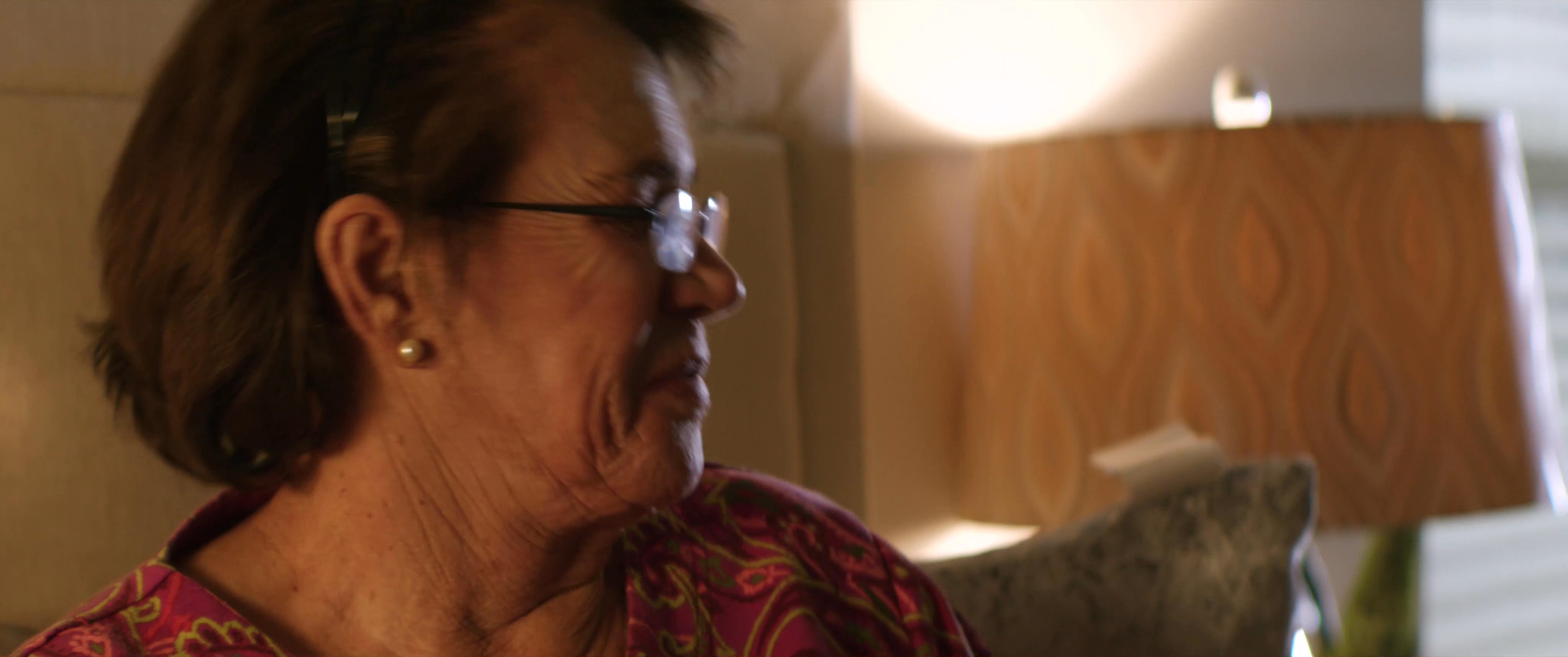 A young man visits an elderly woman in her bedroom, providing assistance and companionship. He hands her a drink while she rests in bed, illustrating a moment of supportive care.