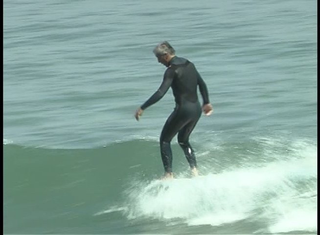 An experienced surfer navigates an ocean wave on a longboard, followed by a transition to an interview with a man wearing a tropical shirt and sunglasses in a garden setting.