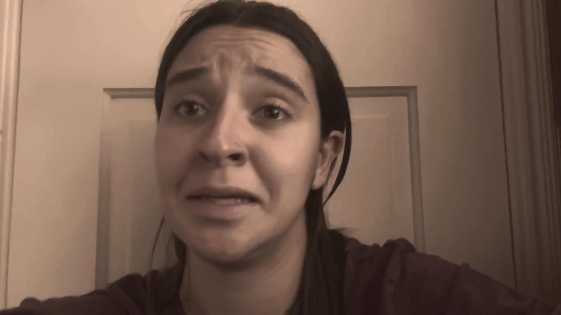 A young woman speaks directly to the camera, sharing a personal narrative while visibly struggling with shifting emotions, ranging from contemplation to visible sadness.