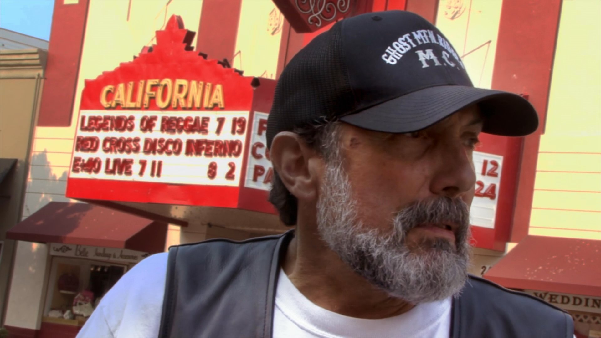 A member of the Ghost Mountain Riders motorcycle club speaks during an interview on a city street, while police officers patrol in the background.