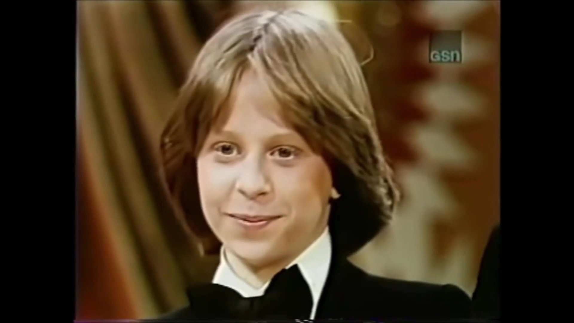 A game show host in a tuxedo engages in a friendly, lighthearted conversation with a young male contestant on a studio stage.