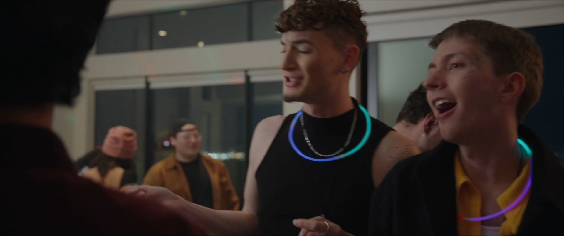 A group of young adults gathers at a brightly lit, modern residence for a lively social event. Participants engage in conversation, share drinks, and embrace as they socialize throughout the evening.