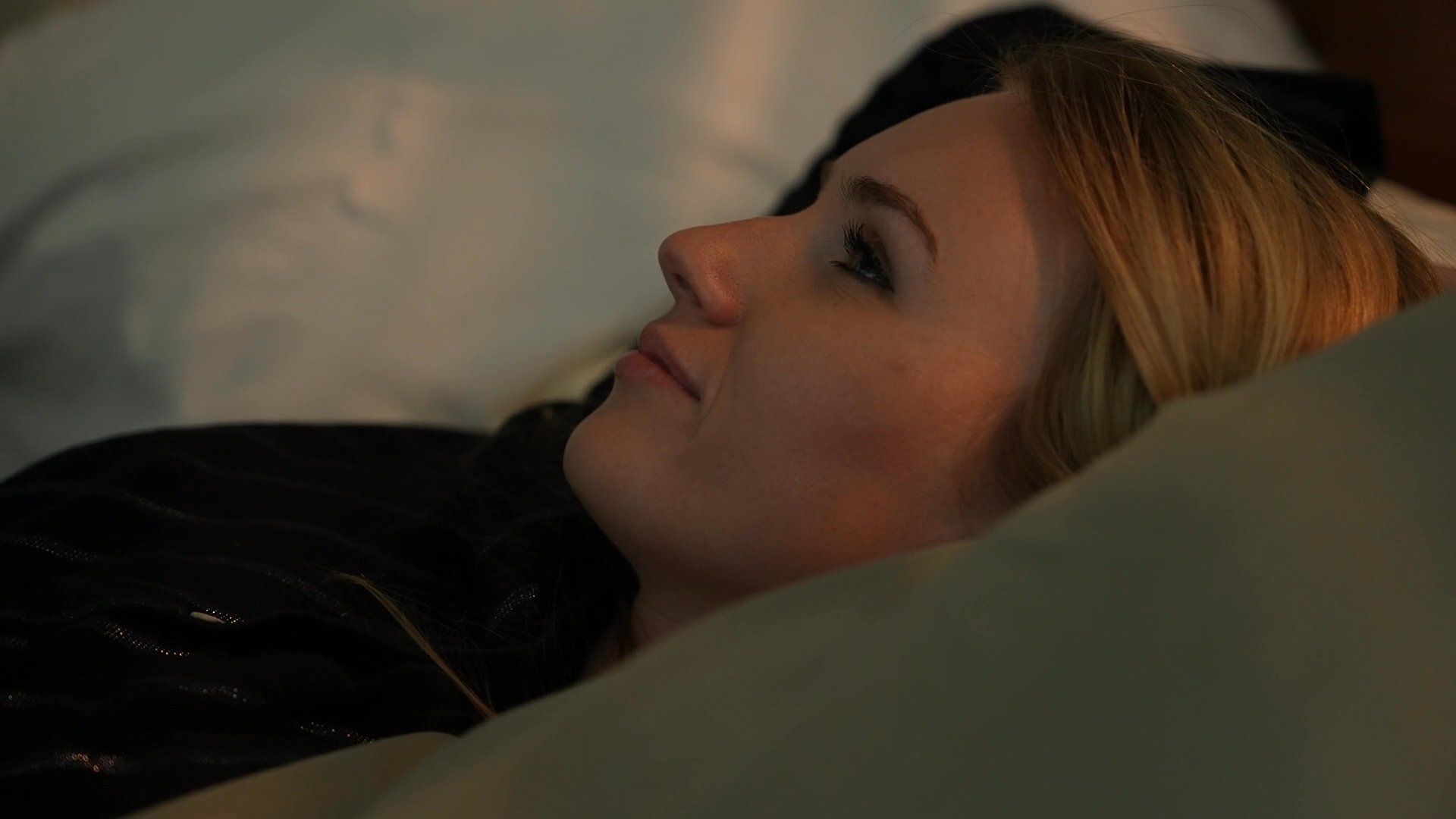A woman wakes from sleep, stretches in bed, and sits up in a dimly lit, cozy bedroom environment.