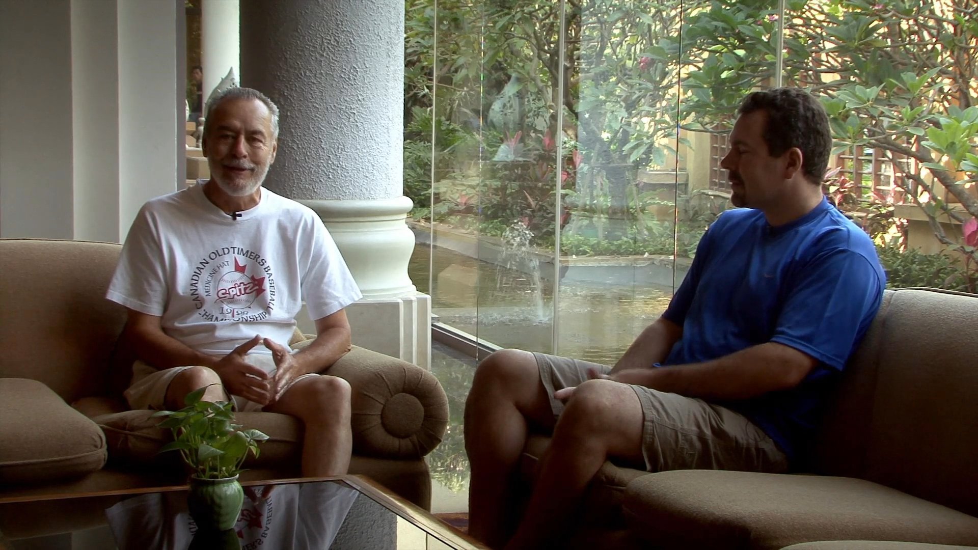 Two men engage in a structured conversation while seated on a sofa in a sunlit, semi-outdoor setting. They exchange perspectives through active listening and verbal communication.