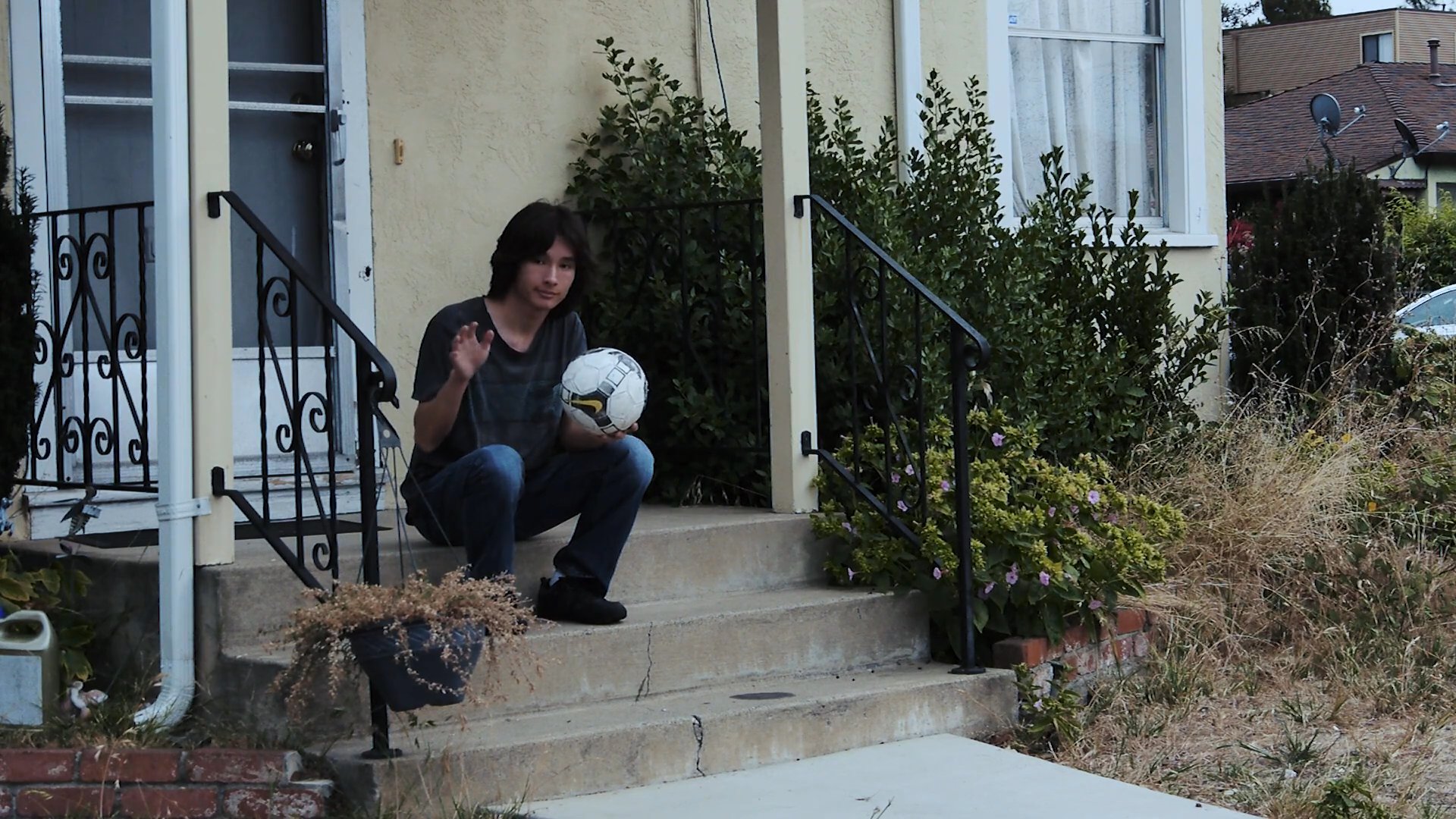 A woman operates a car through a residential area while engaging with someone waiting on a porch with a soccer ball. She eventually proceeds to drive away from the scene.