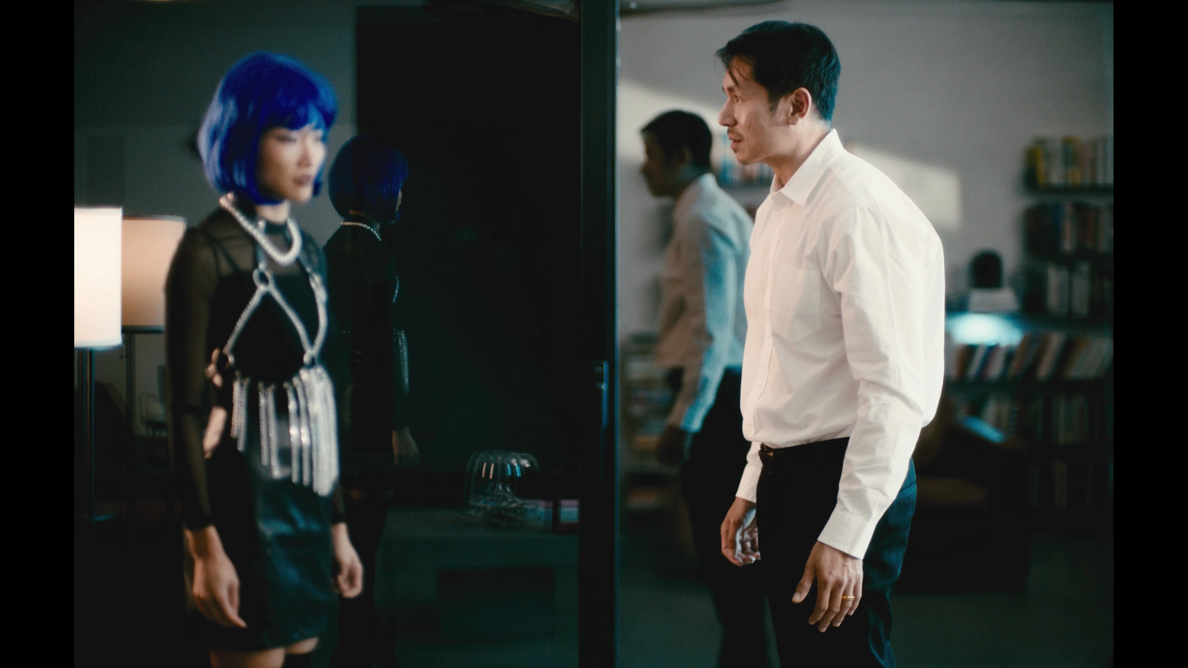A woman with blue hair and a man in a white dress shirt engage in a serious, tense conversation within a residential living room.