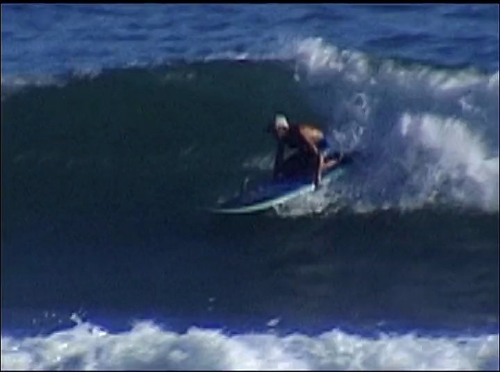 A surfer maneuvers a longboard along the face of a breaking ocean wave. The sequence captures the transition from the wave cresting to a controlled ride across the open water.