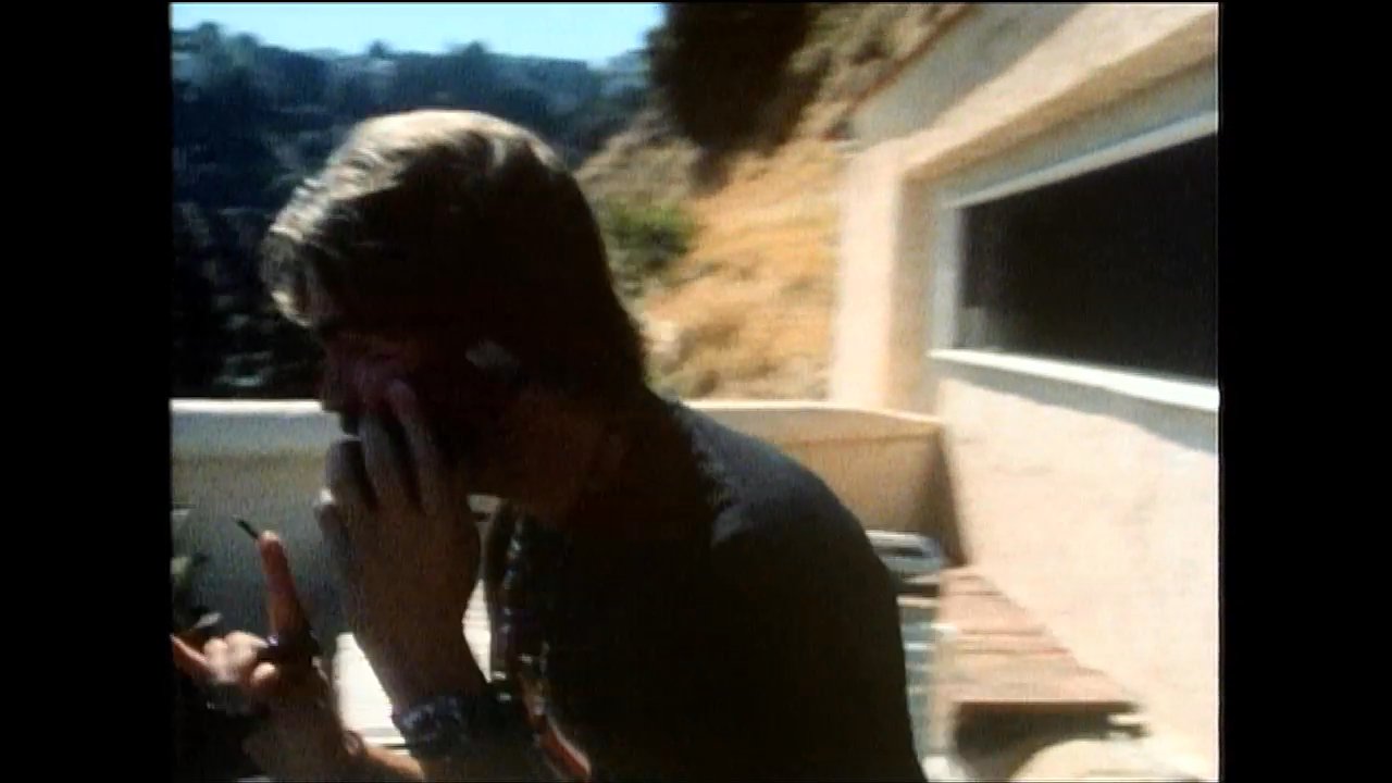 A woman relaxes on a rooftop with cucumber slices on her eyes while a man stands nearby engaged in a phone call overlooking a cityscape.