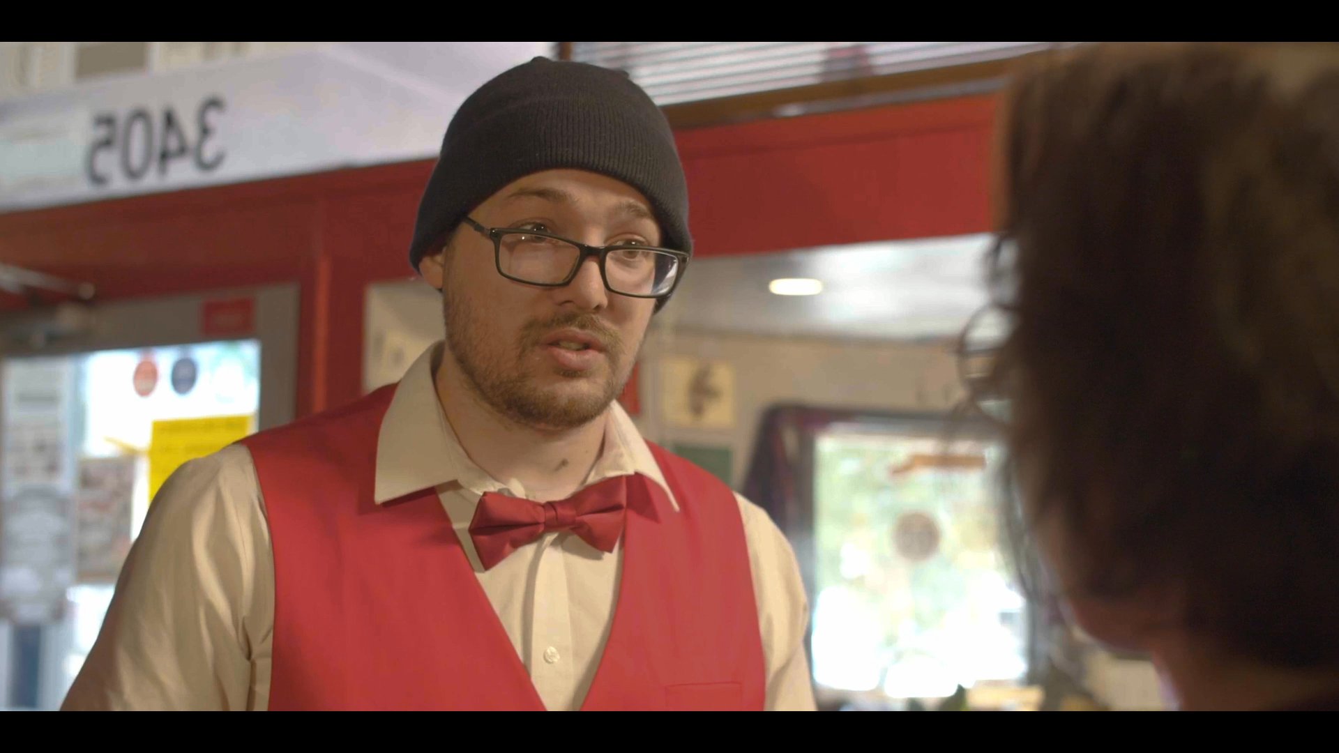 A theatre concession worker in uniform discusses details with a patron in a dimly lit lobby. The two engage in an earnest conversation near a popcorn machine.