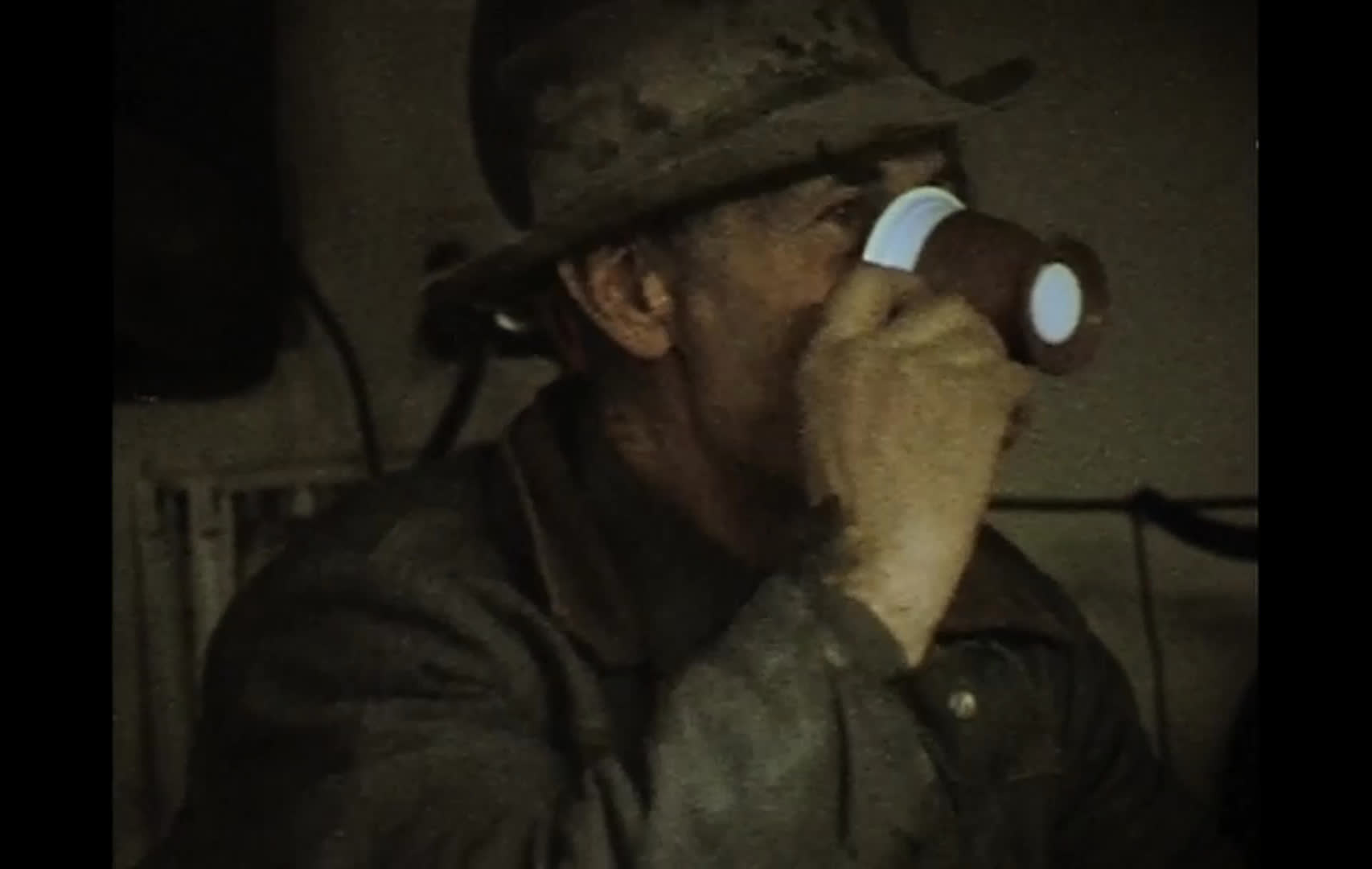 In a dim, rustic interior, a woman and a man in a cap talk beside bottles while an older man in a wide-brimmed hat drinks from a paper cup. The group shares a relaxed, humorous exchange about whiskey, coffee, and a donut.
