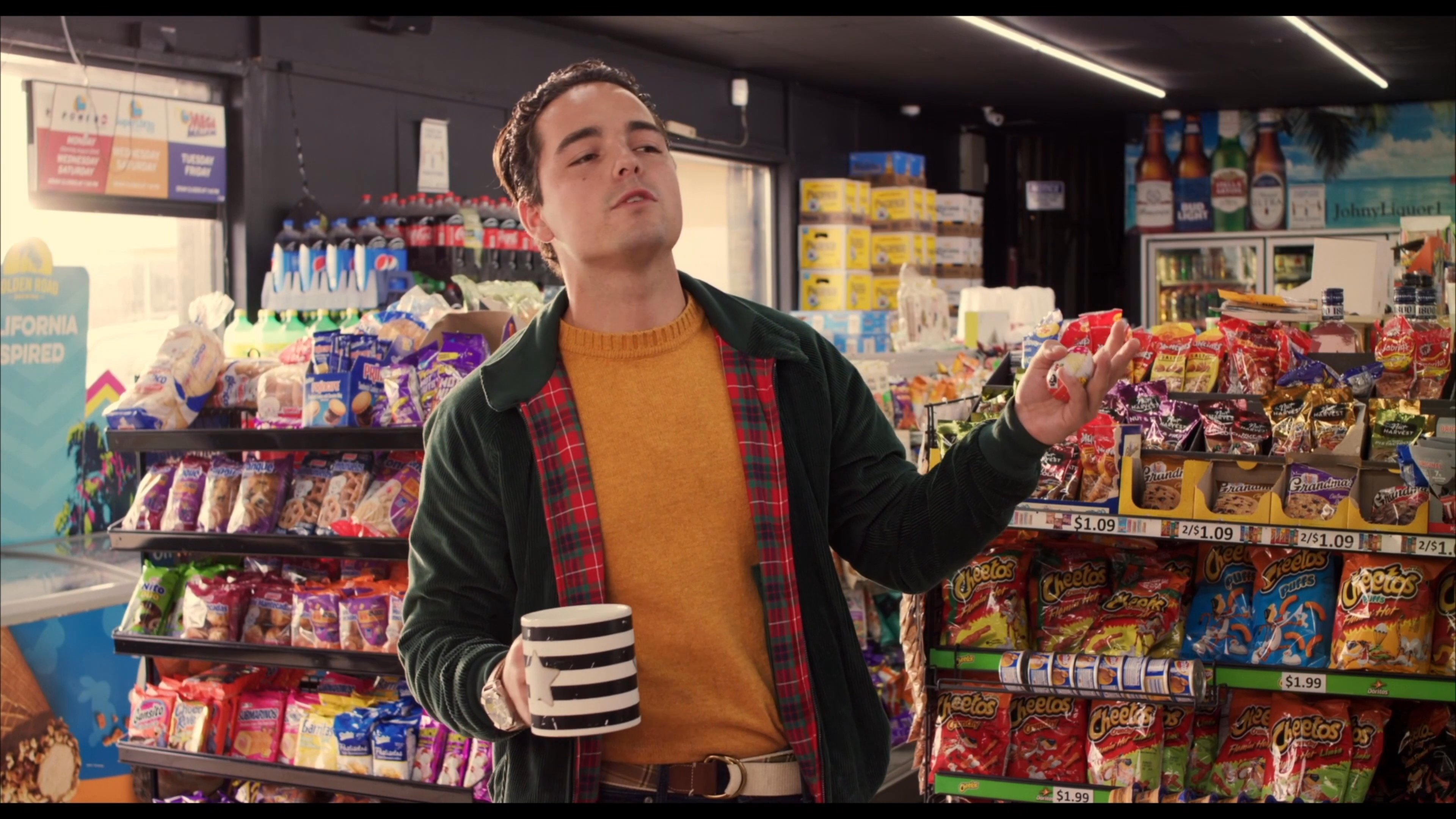 A customer holding a coffee mug engages in conversation while browsing a convenience store aisle, while a nearby employee looks through a magazine.