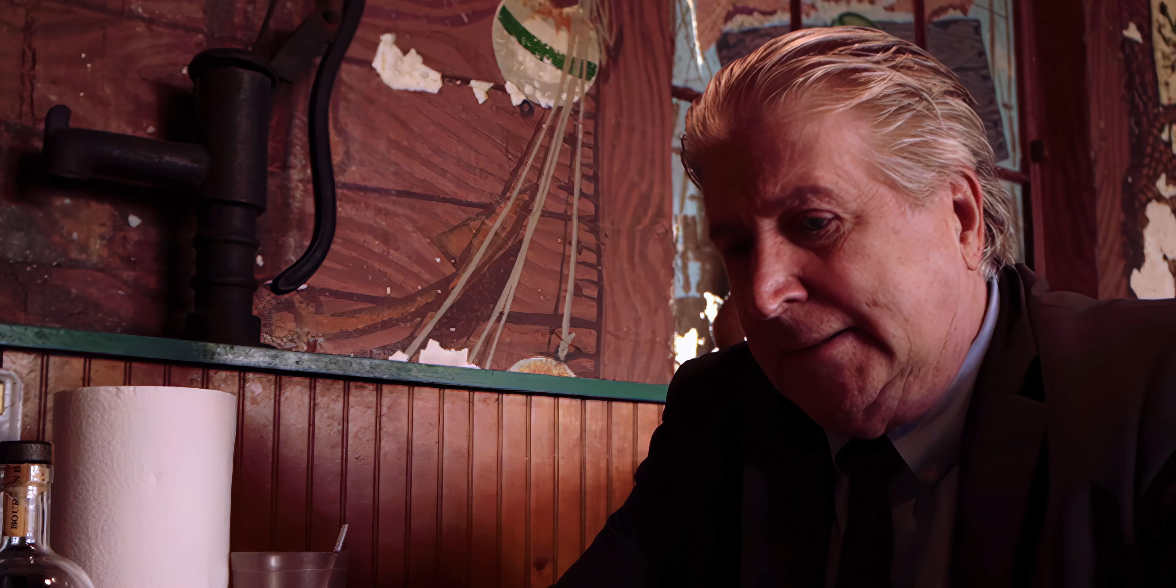 Two men engage in a serious, focused conversation while seated inside a vintage-themed diner. The individual in the suit gestures expressively as he speaks to his companion.