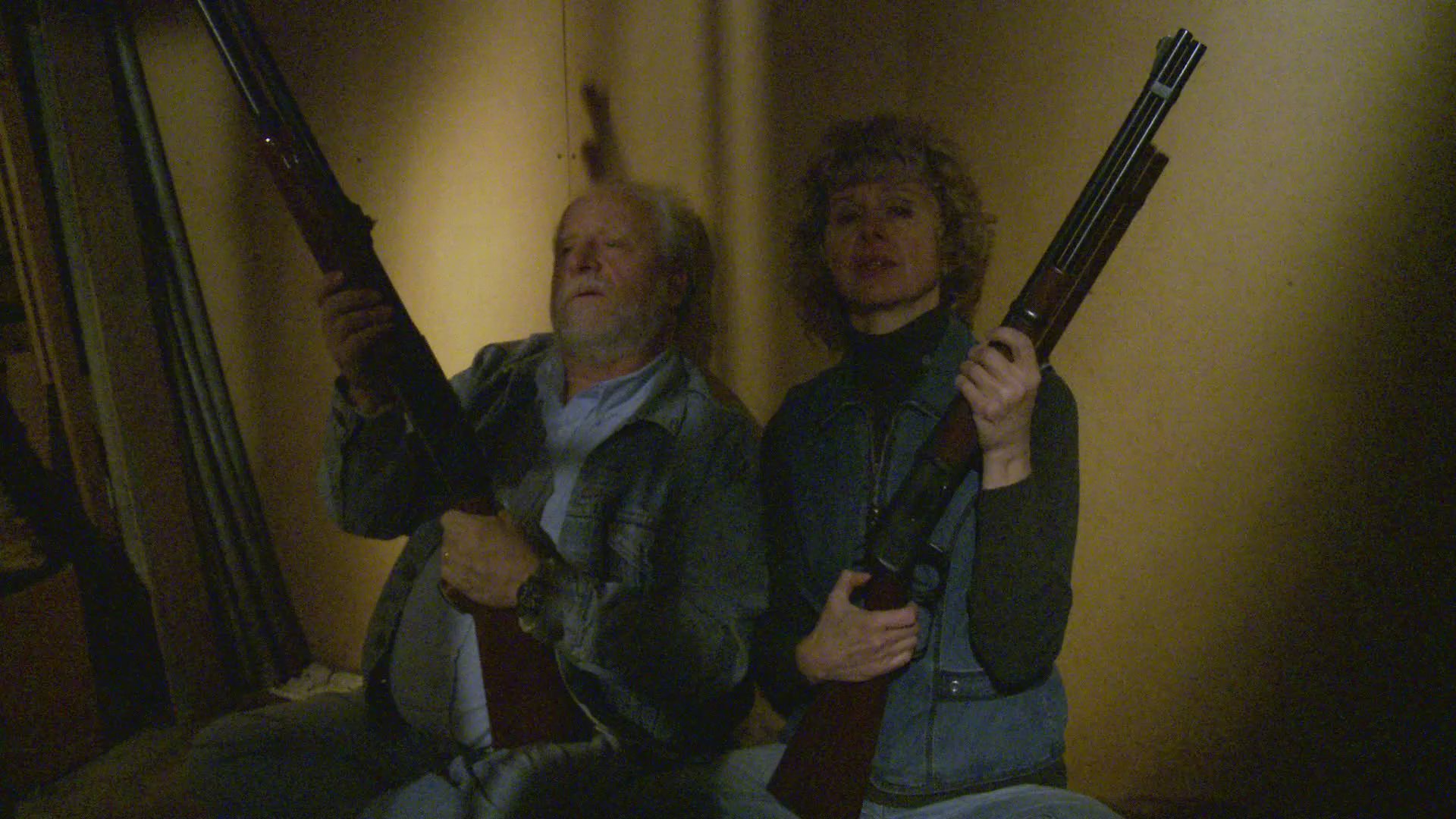 An older man and woman wait vigilantly with shotguns in a dim room before observing an intruder attempting to enter their residence through a door.