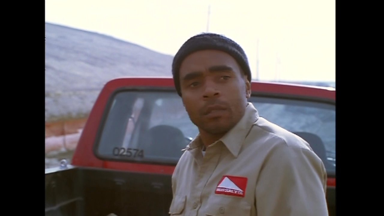Two utility workers in uniform coordinate tasks beside a red pickup truck parked on a desolate dirt path. After a brief interaction, one worker walks away into the expansive, barren landscape.