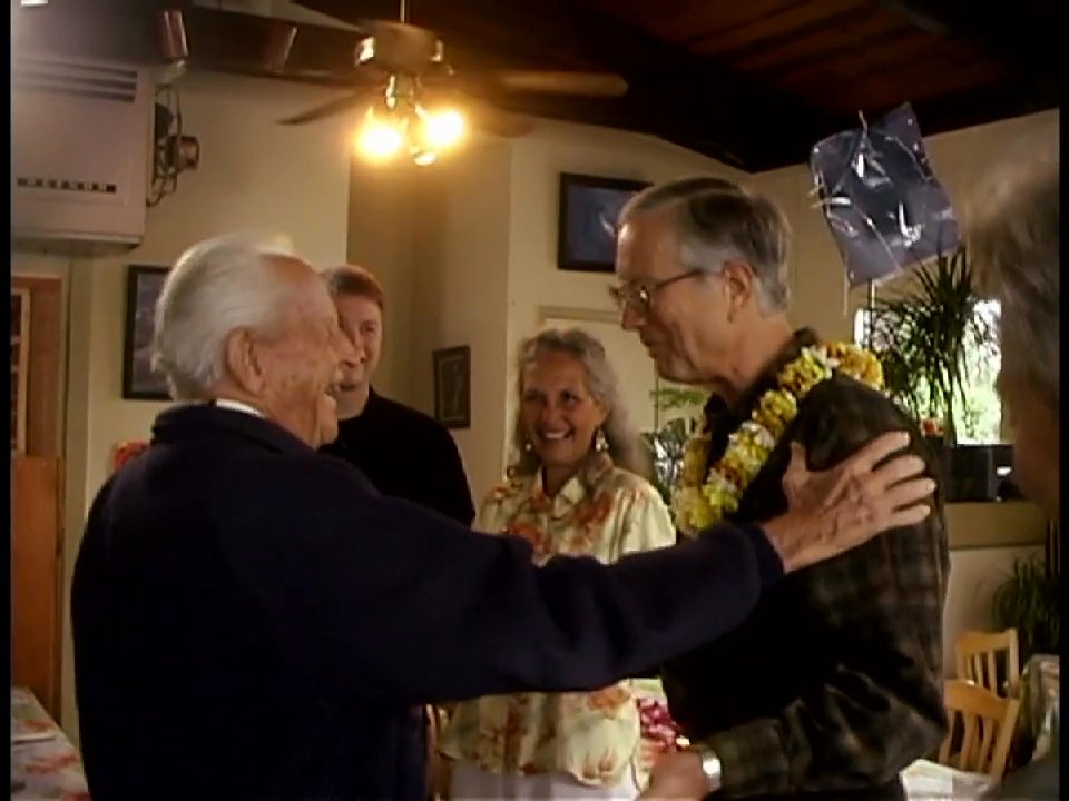 Two senior men greet each other warmly with handshakes and smiles in a room decorated with surfboards. A woman in a floral shirt observes the celebratory reunion in the background.
