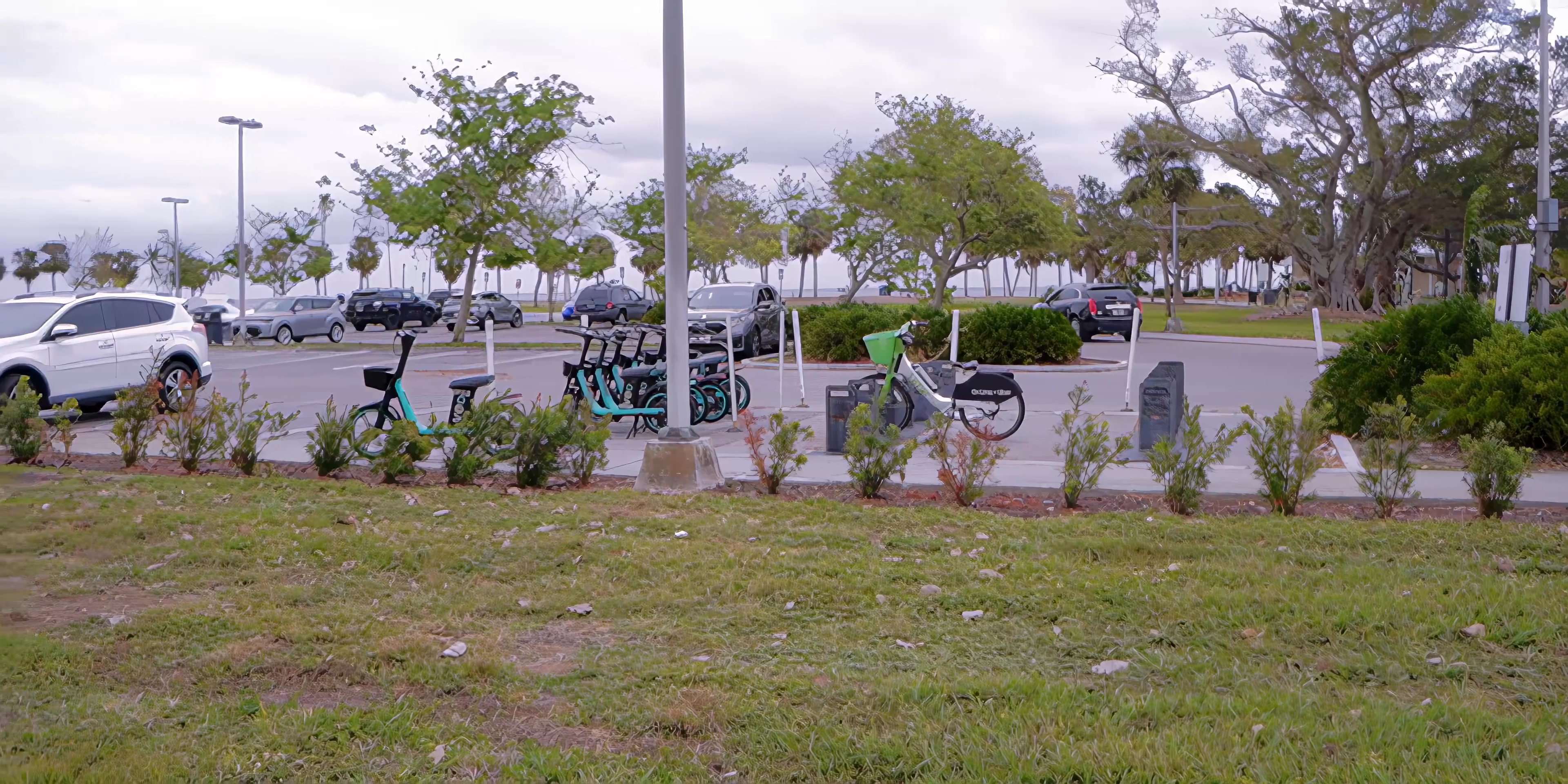 A black SUV drives along a coastal road past a public park with rental bicycles before the perspective shifts to two men walking toward the ocean shoreline.