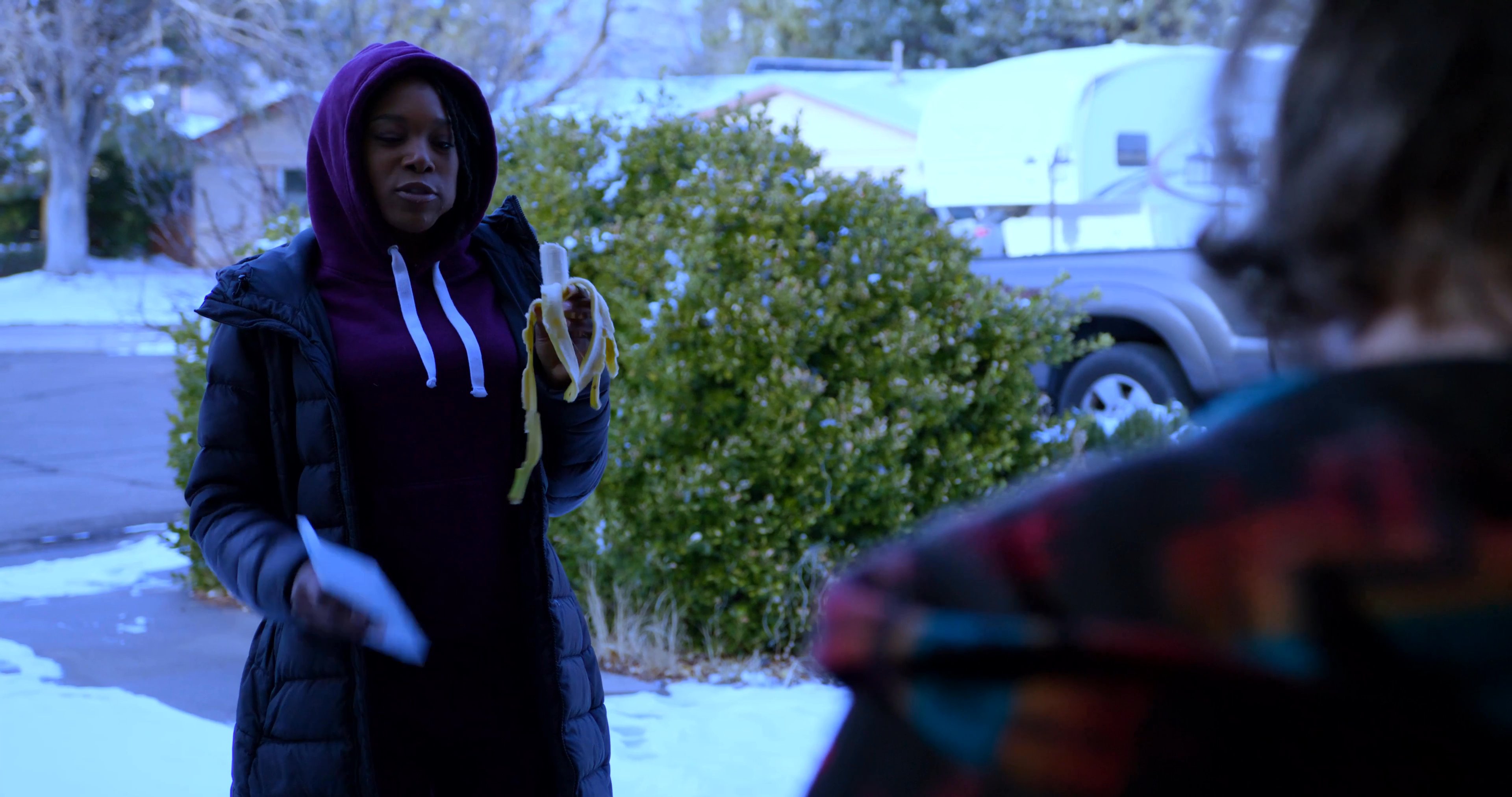 A man in a patterned robe interacts with a woman in a hooded jacket in a residential driveway setting. The encounter involves a brief, casual exchange between the two individuals.