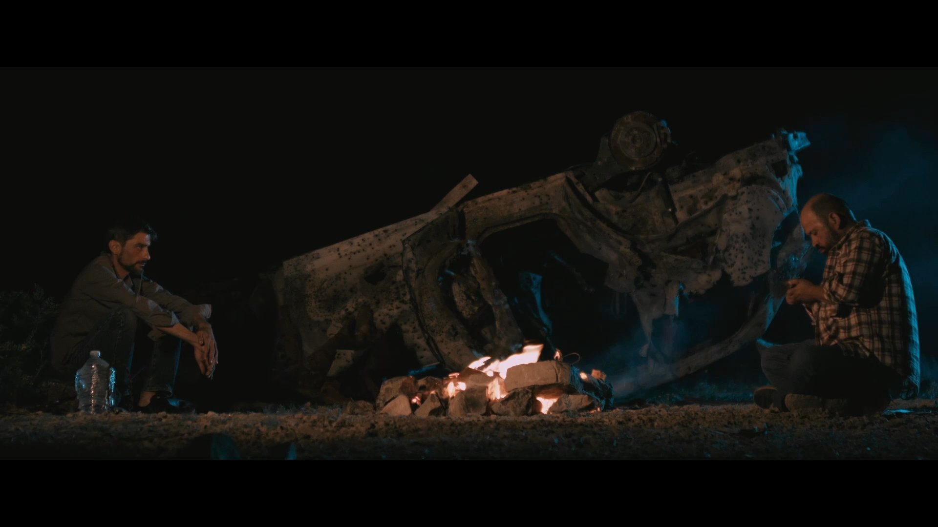 Two individuals engage in a quiet dialogue beside a campfire in a dark, outdoor setting while one prepares tinder for the fire.