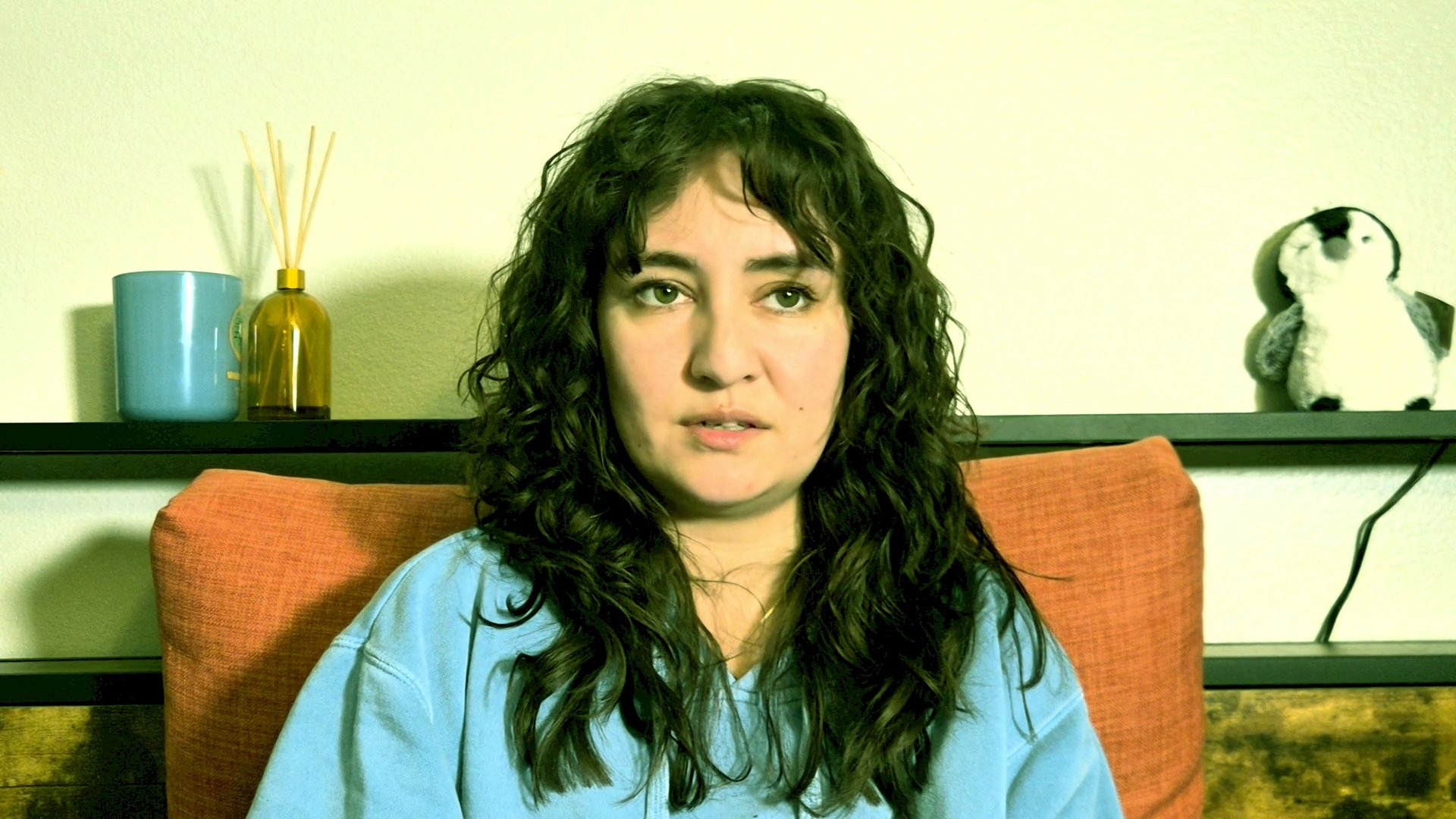 A woman speaks directly to the camera, sharing a narrative in an intimate indoor setting. Her expressions shift between reflection and engagement as she articulates her thoughts.