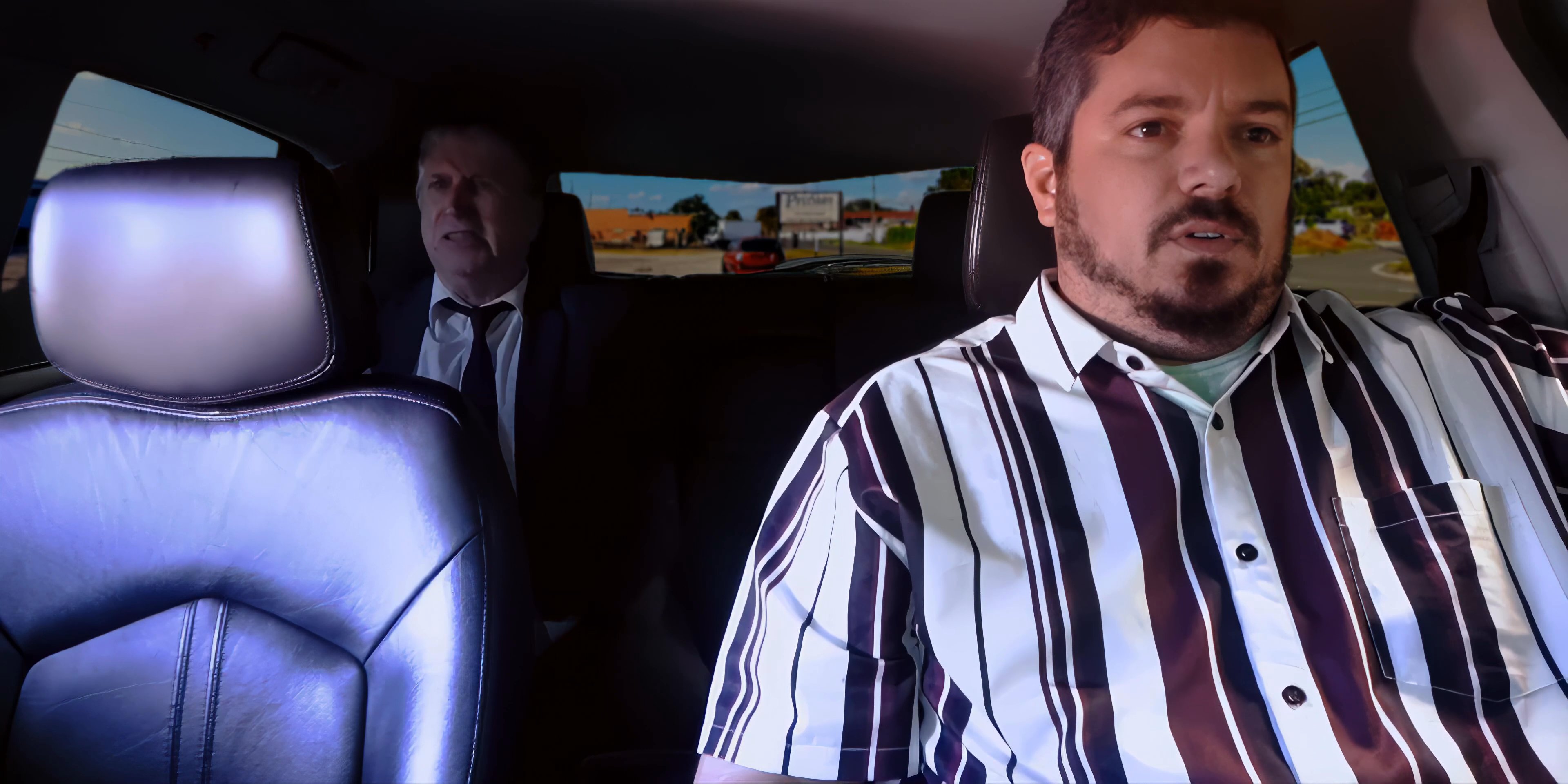 A man drives a vehicle while engaging in a serious discussion with a passenger seated in the back. The scene focuses on their ongoing interaction during a daytime commute.