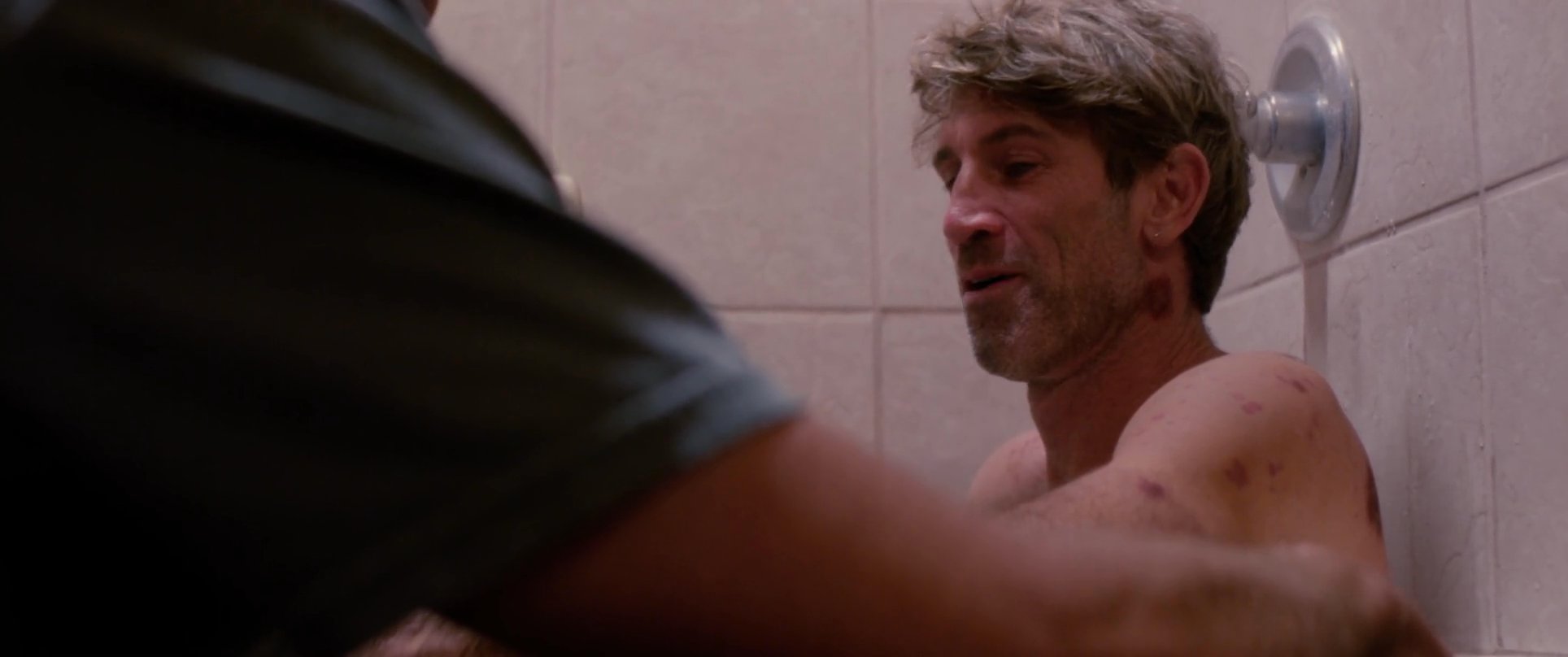 A man provides physical assistance to another man in a shower setting, highlighting a moment of personal care and vulnerability.