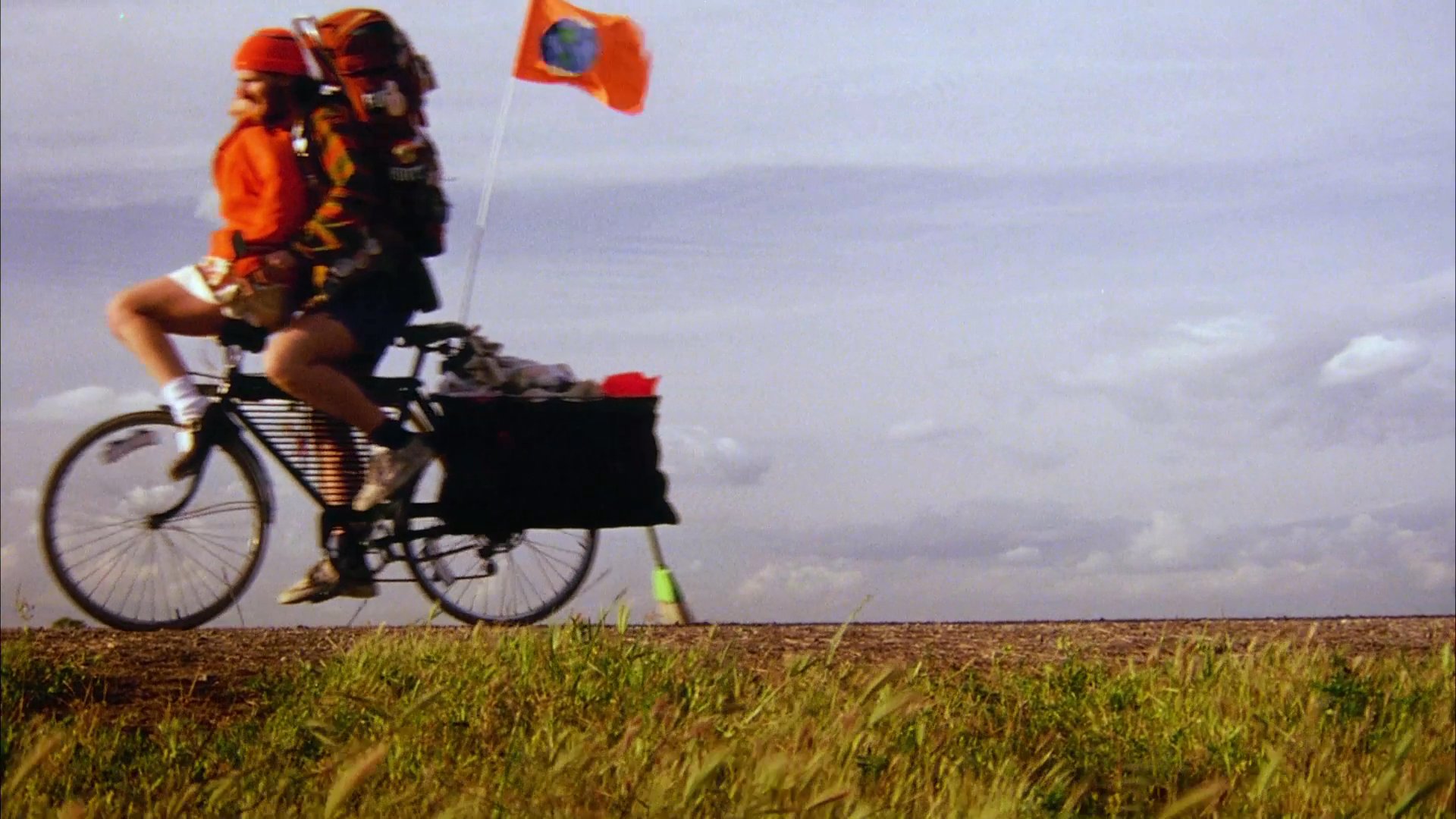 A couple travels together on a single bicycle through a rural landscape. They pedal along an open road, carrying heavy luggage and gear on their journey.