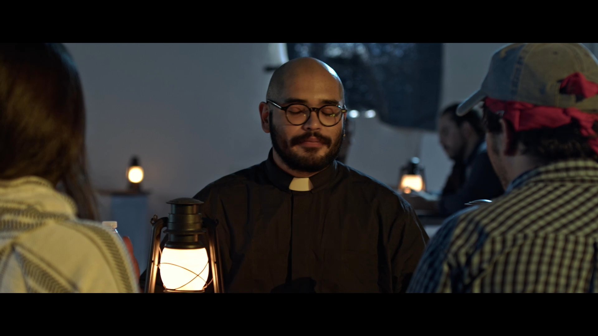 A priest converses with travelers in a dimly lit room, providing information about their journey from Juarez. The exchange occurs under the warm light of a lantern as the group seeks rest for the night.