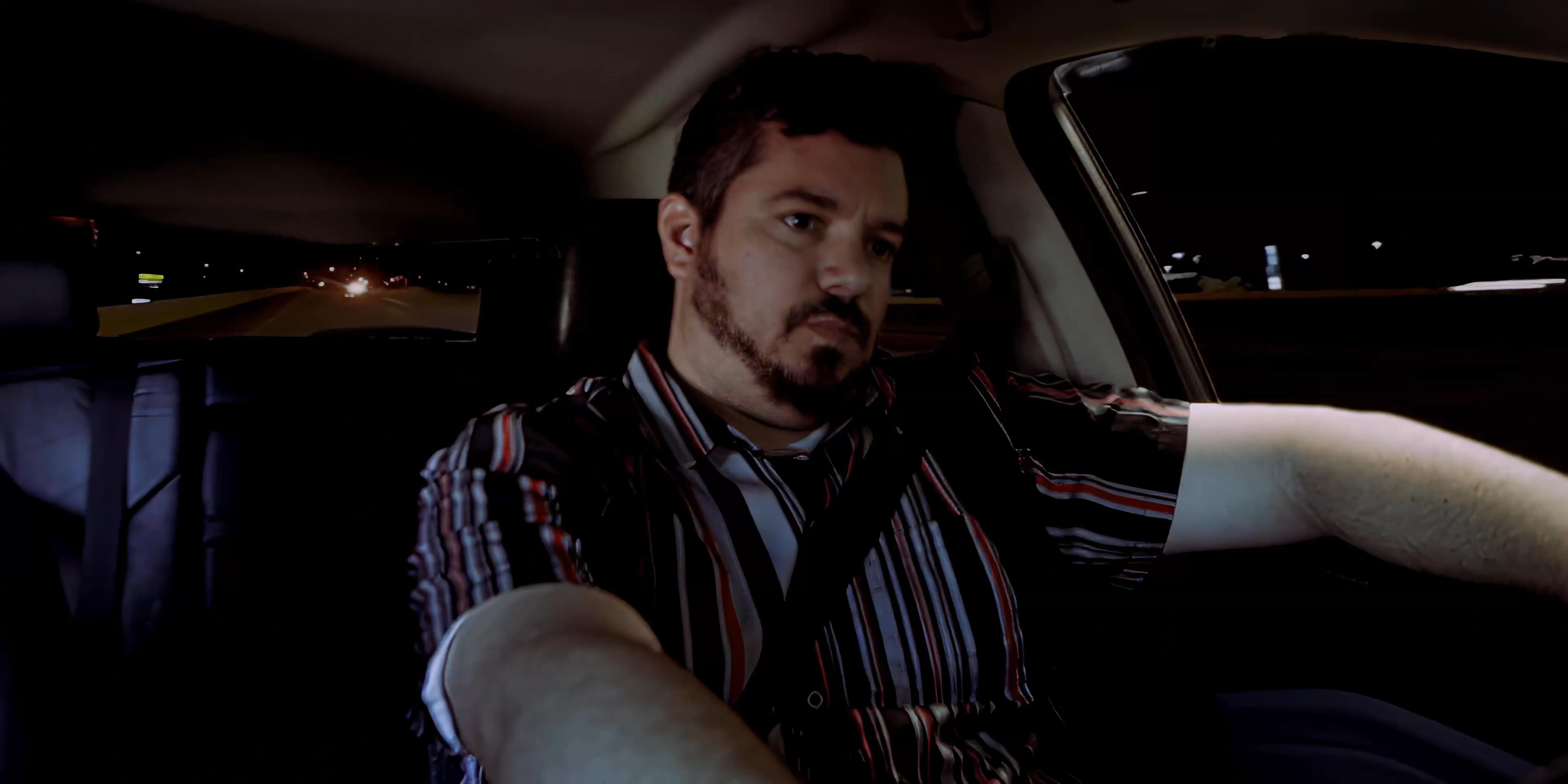 A man eats a meal inside a vehicle at night before driving across a city with an illuminated skyline. The scene captures a solitary evening commute through an urban environment.