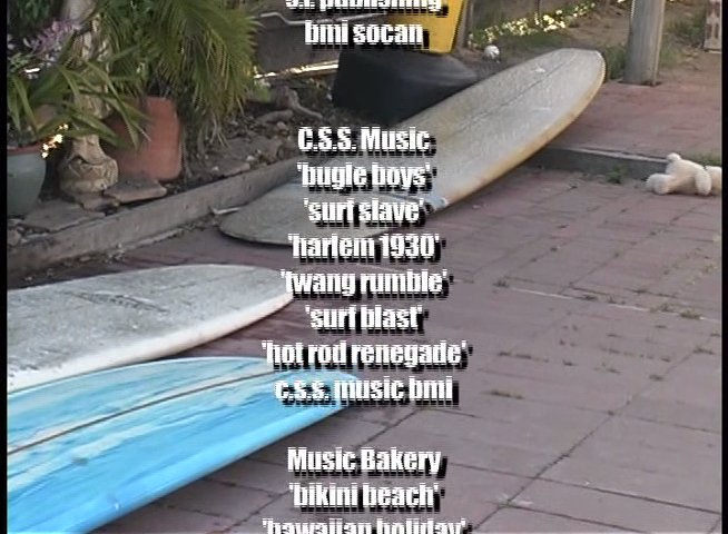 A surfer carries a longboard near a beach house while end credits roll over footage of ocean waves and surf culture.