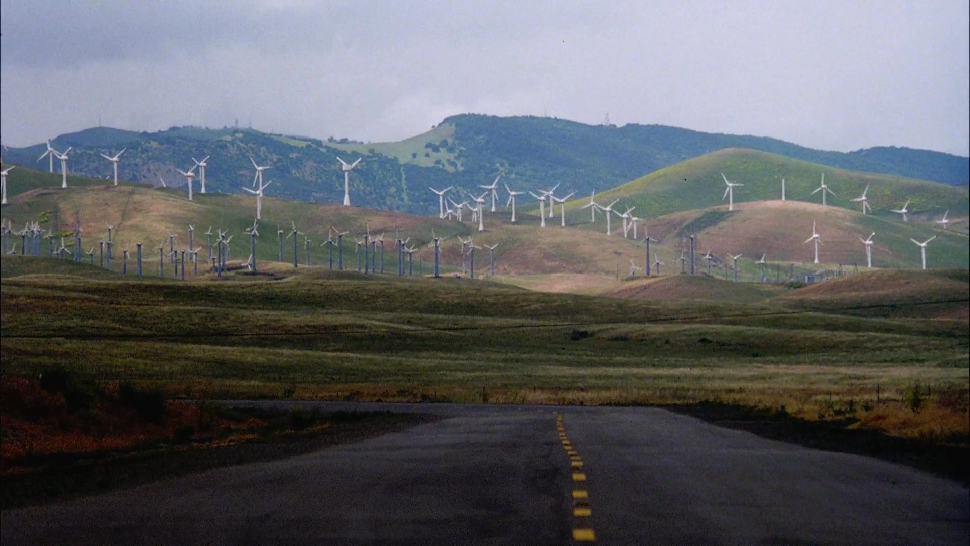 A traveler equipped with a large backpack and bicycle prepares for a journey through a vast, rural landscape populated by wind turbines. The sequence captures a contemplative moment before the traveler continues their outdoor expedition.