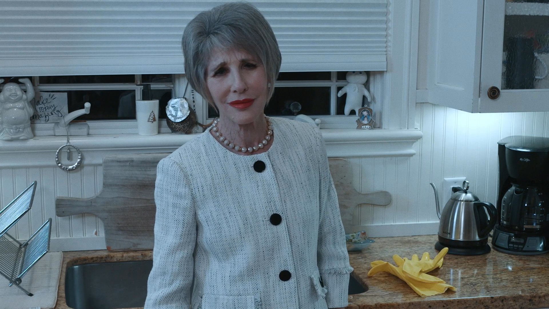 A man engages in a tense conversation with an older woman in a kitchen setting. The exchange highlights a dynamic of interpersonal conflict through their expressive gestures and focused attention.