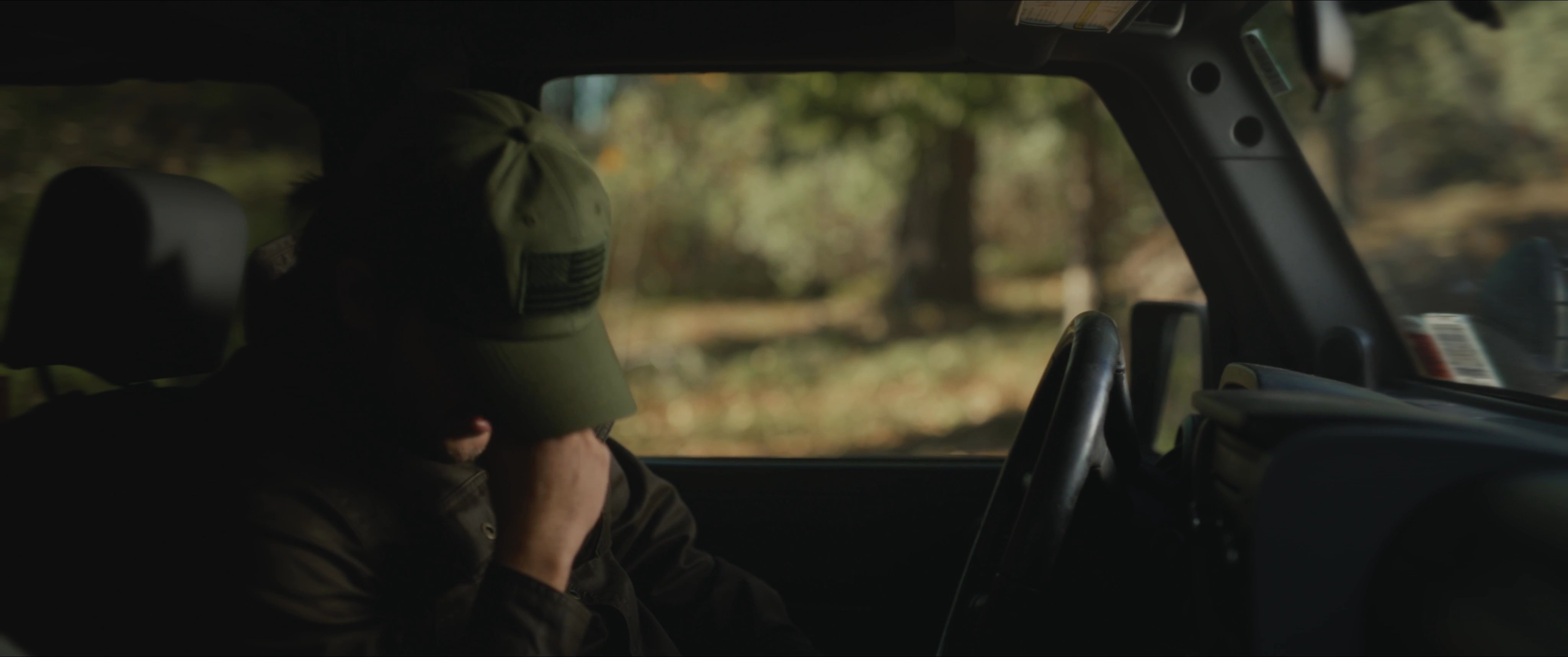 A sheriff investigates a scene in a wooded area before returning to his vehicle, where he experiences an intense moment of personal distress.